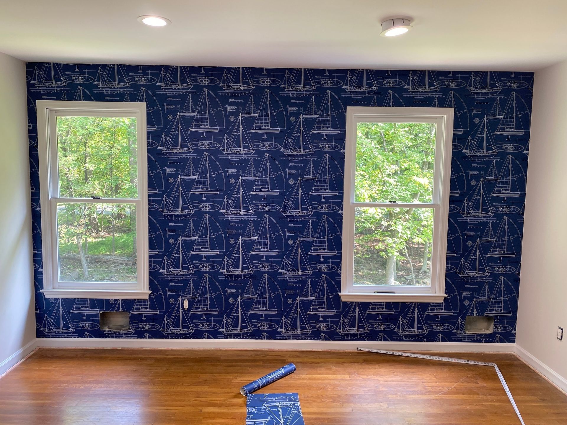 Navy wallpaper with white sailboat pattern between two windows in a room.