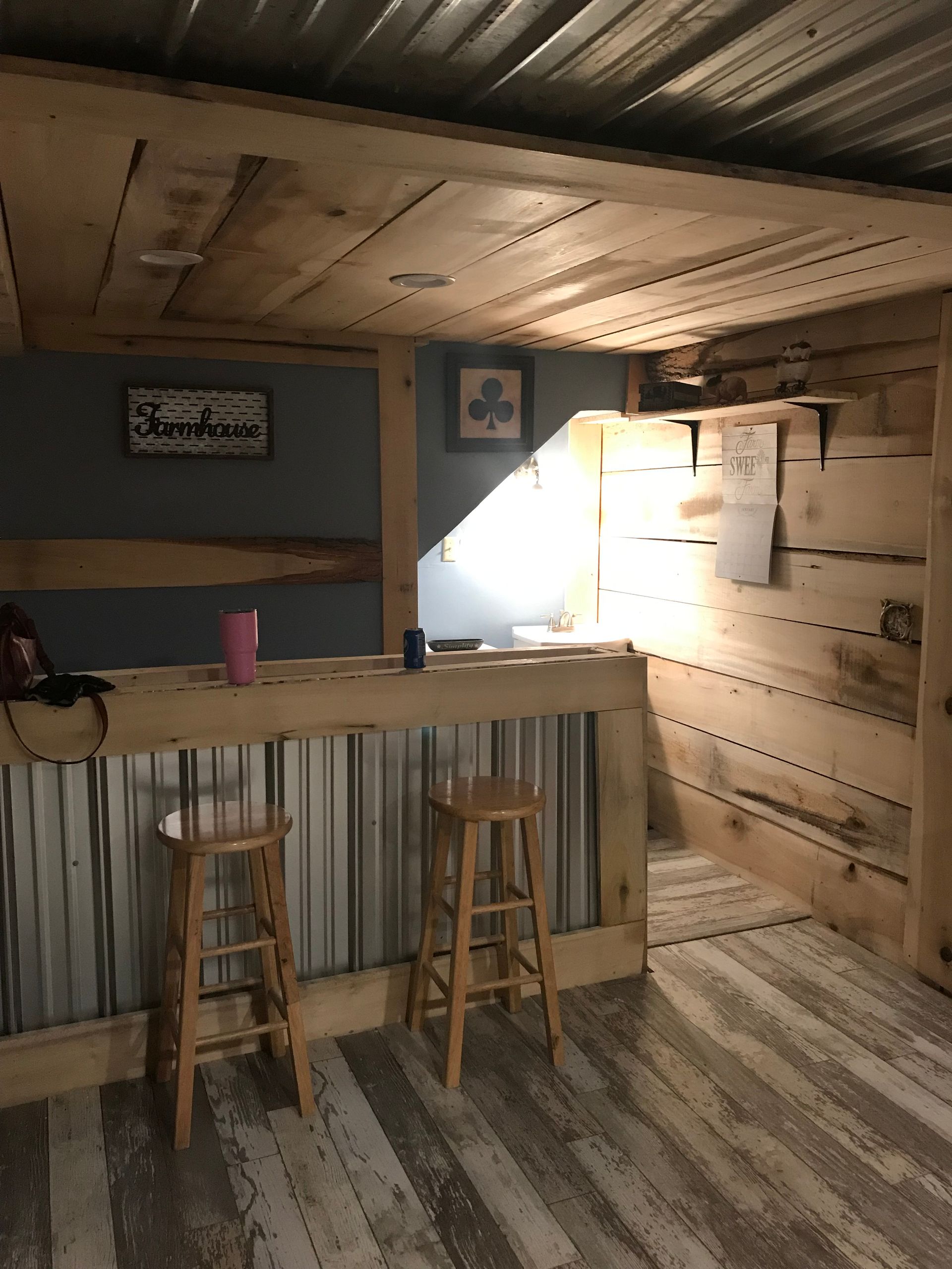 A rustic home bar with wooden paneling, a corrugated metal front, two stools, and a sign on a gray-painted wall.