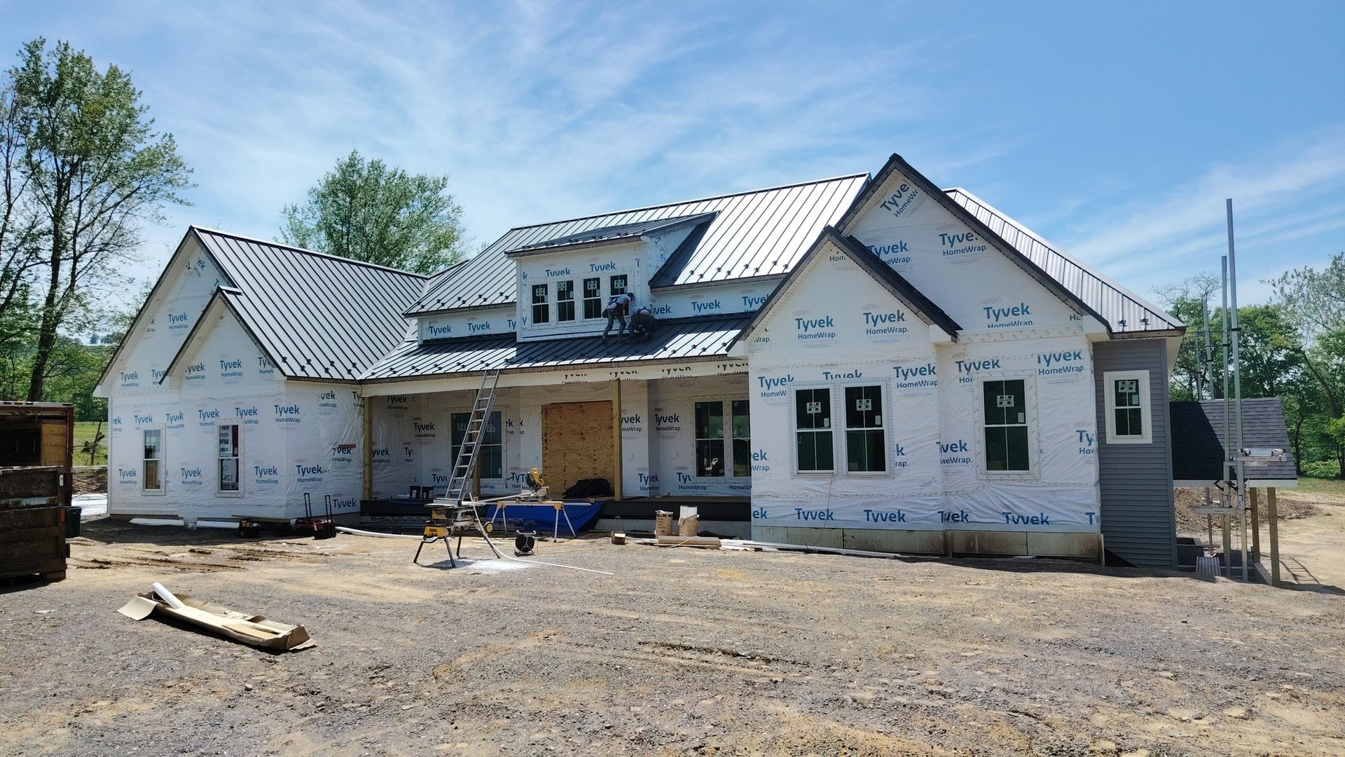 A residential house under construction, with white exterior weather barrier siding, a gray metal roof, and blue sky.