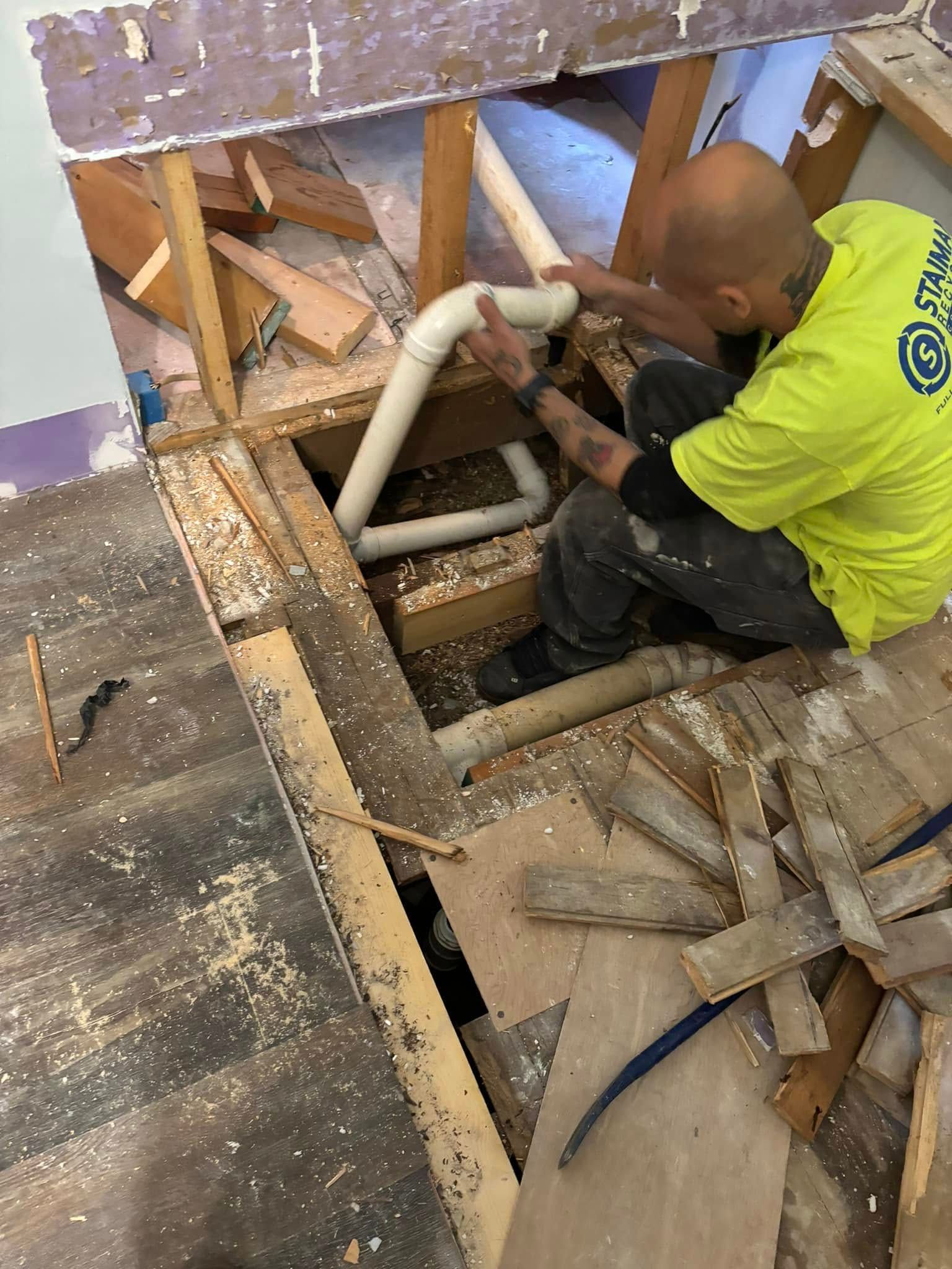 A worker in a bright yellow shirt installs white PVC plumbing pipes beneath an open floor in a construction setting.