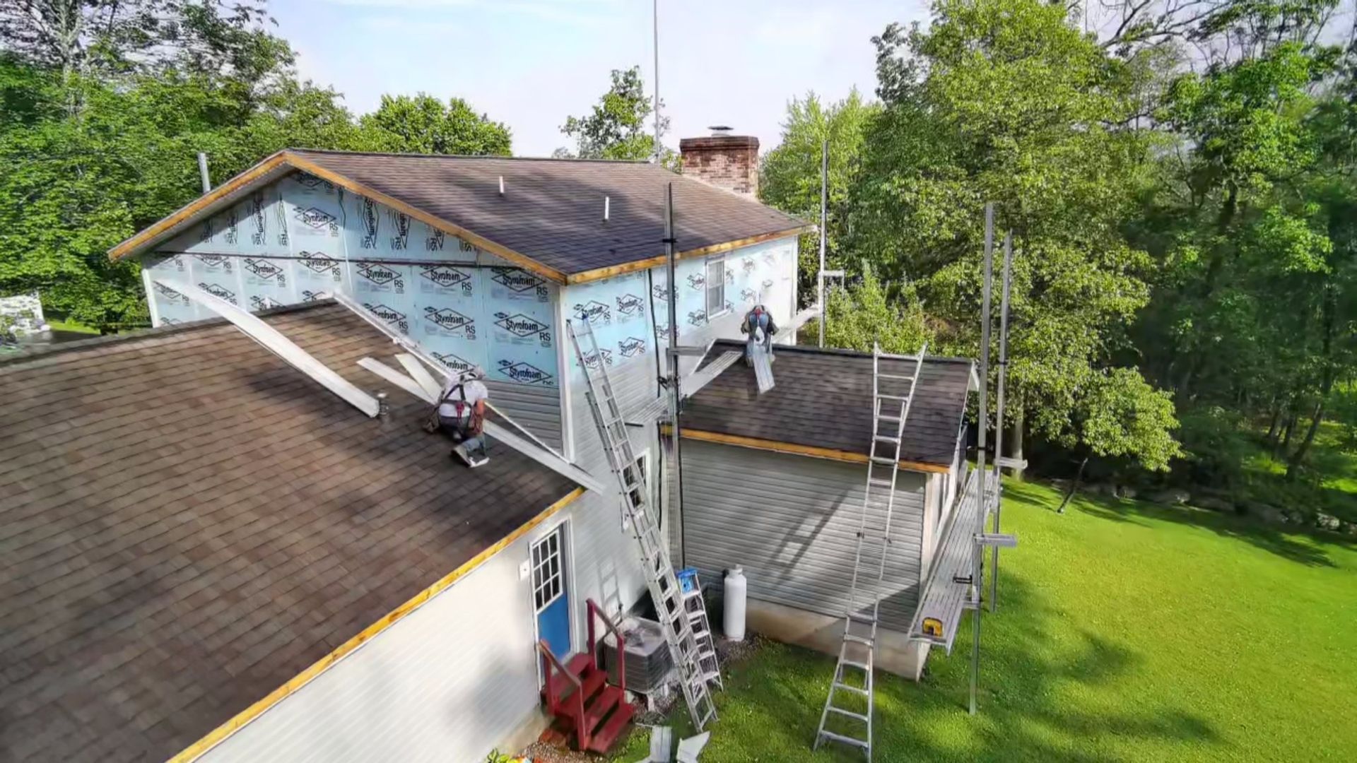 Workers on ladders and roofs perform exterior construction on a two-story home with blue insulation and green trees.
