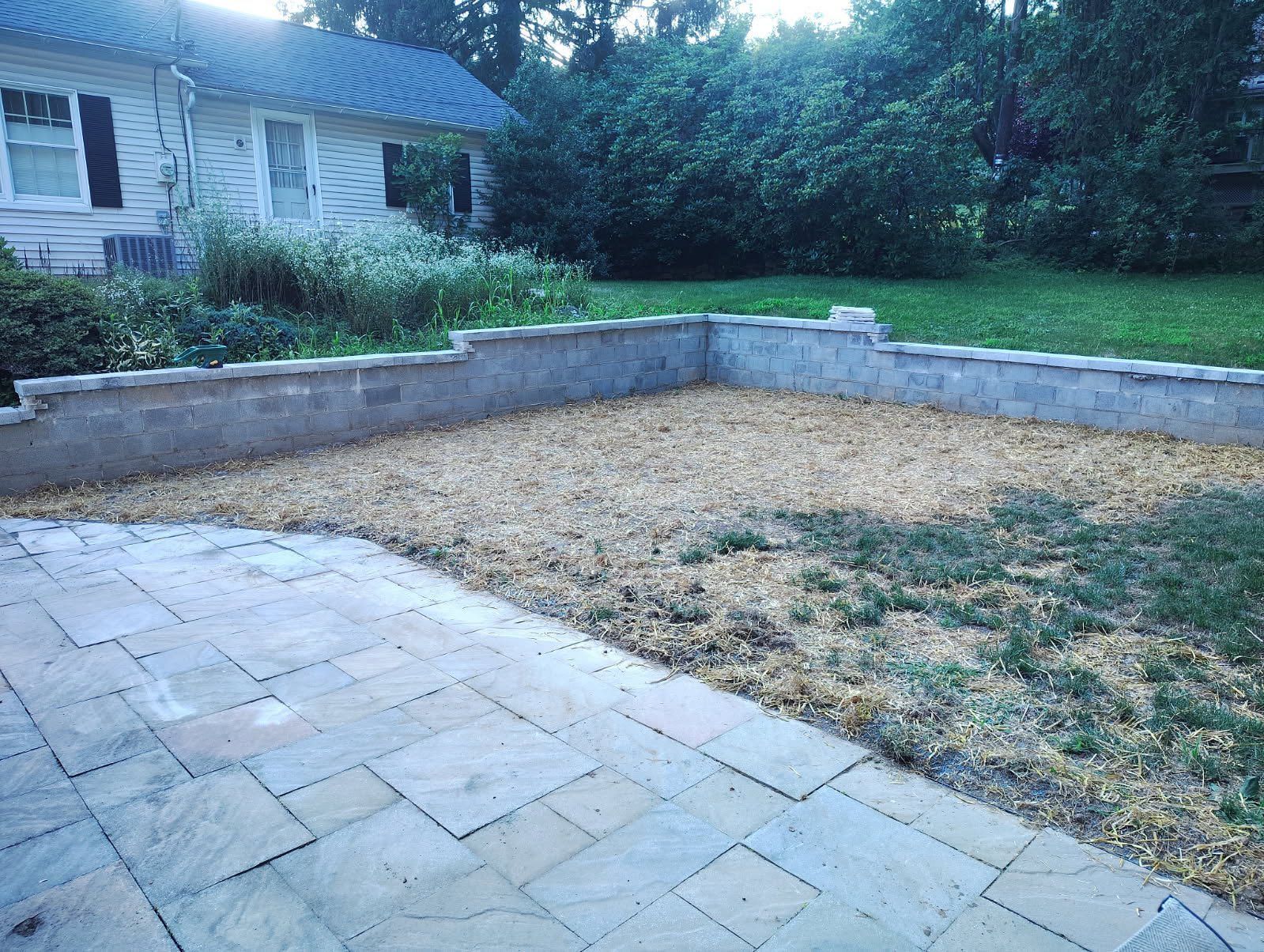 A backyard view featuring a light stone patio in the foreground, a wood-chipped area, and a gray stone retaining wall.