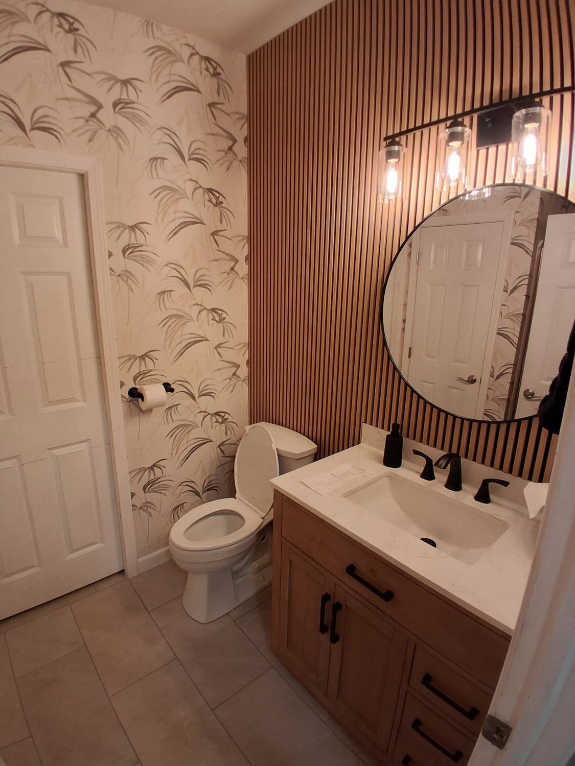 A bathroom with patterned wallpaper, a wooden vanity with a white sink, a round mirror, and a toilet.