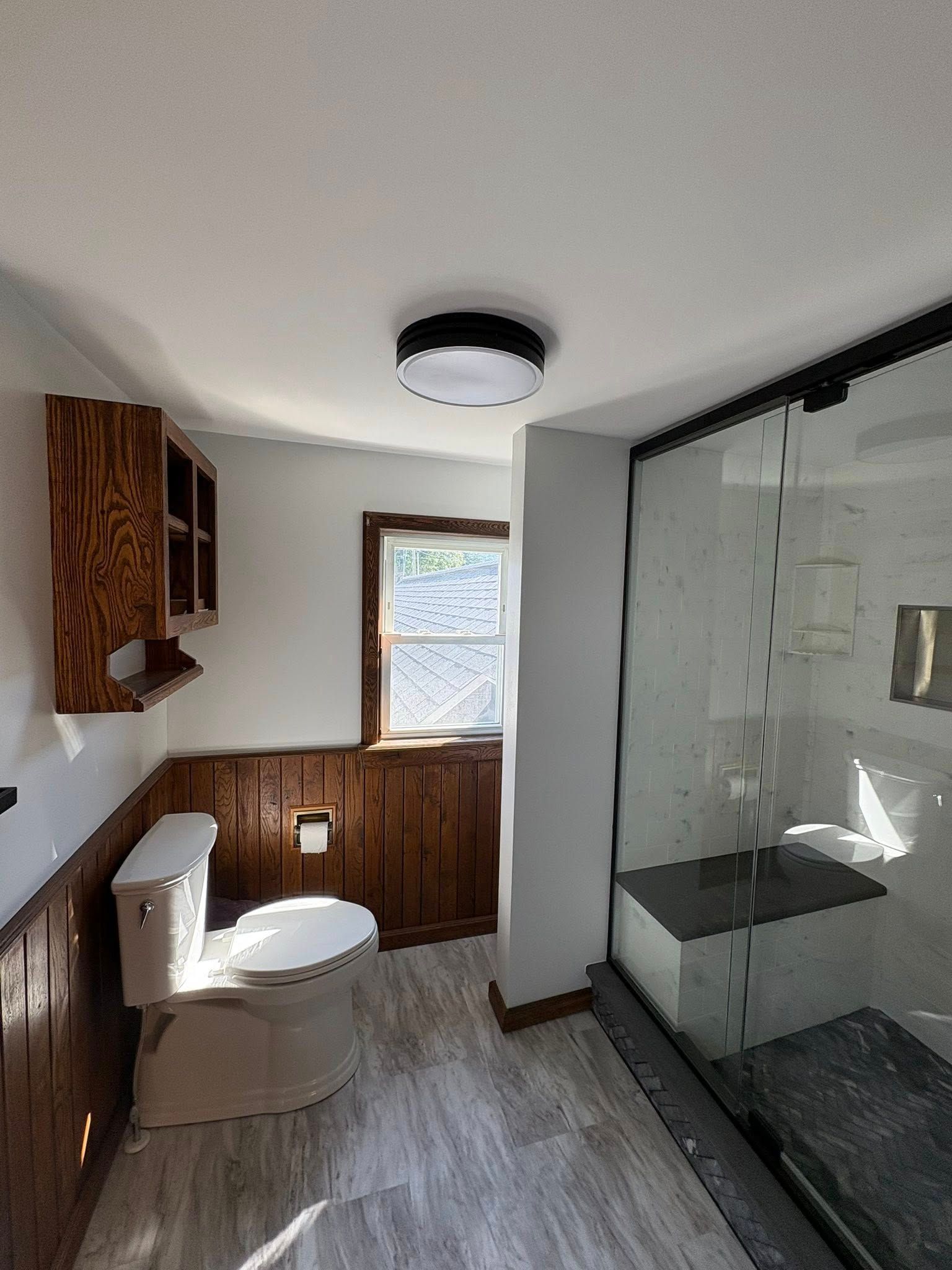 A bathroom with wood paneling, a toilet, a wall-mounted cabinet, a window, and a glass-enclosed shower with a bench.