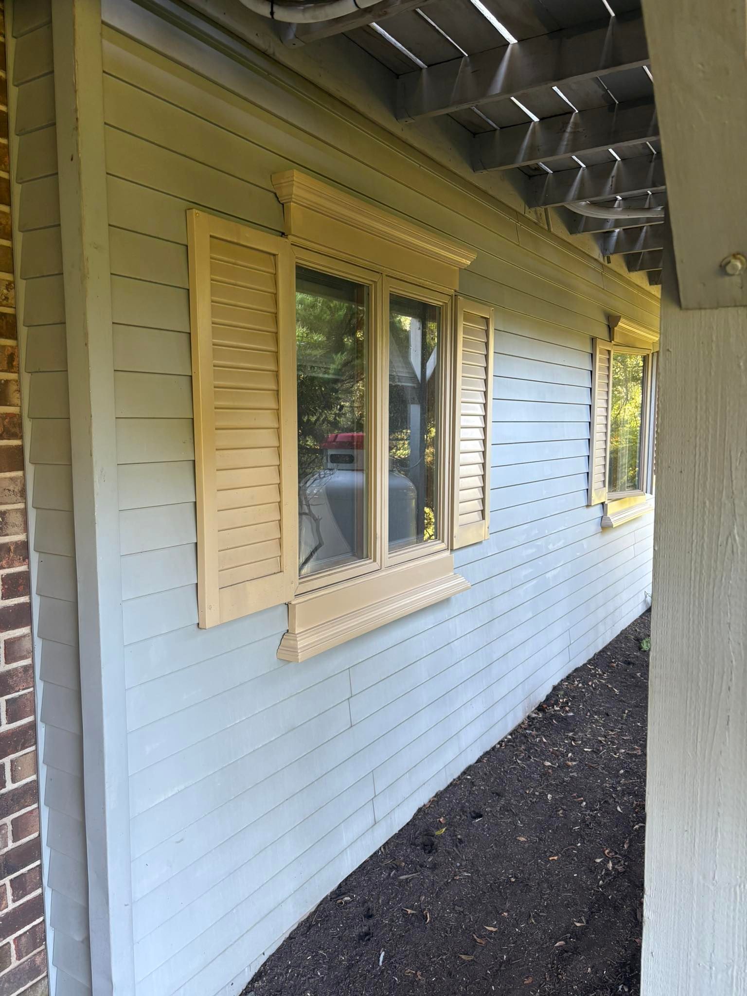 Light grey exterior wall of a house with a window featuring decorative yellow trim, shutters, and a garden bed below.