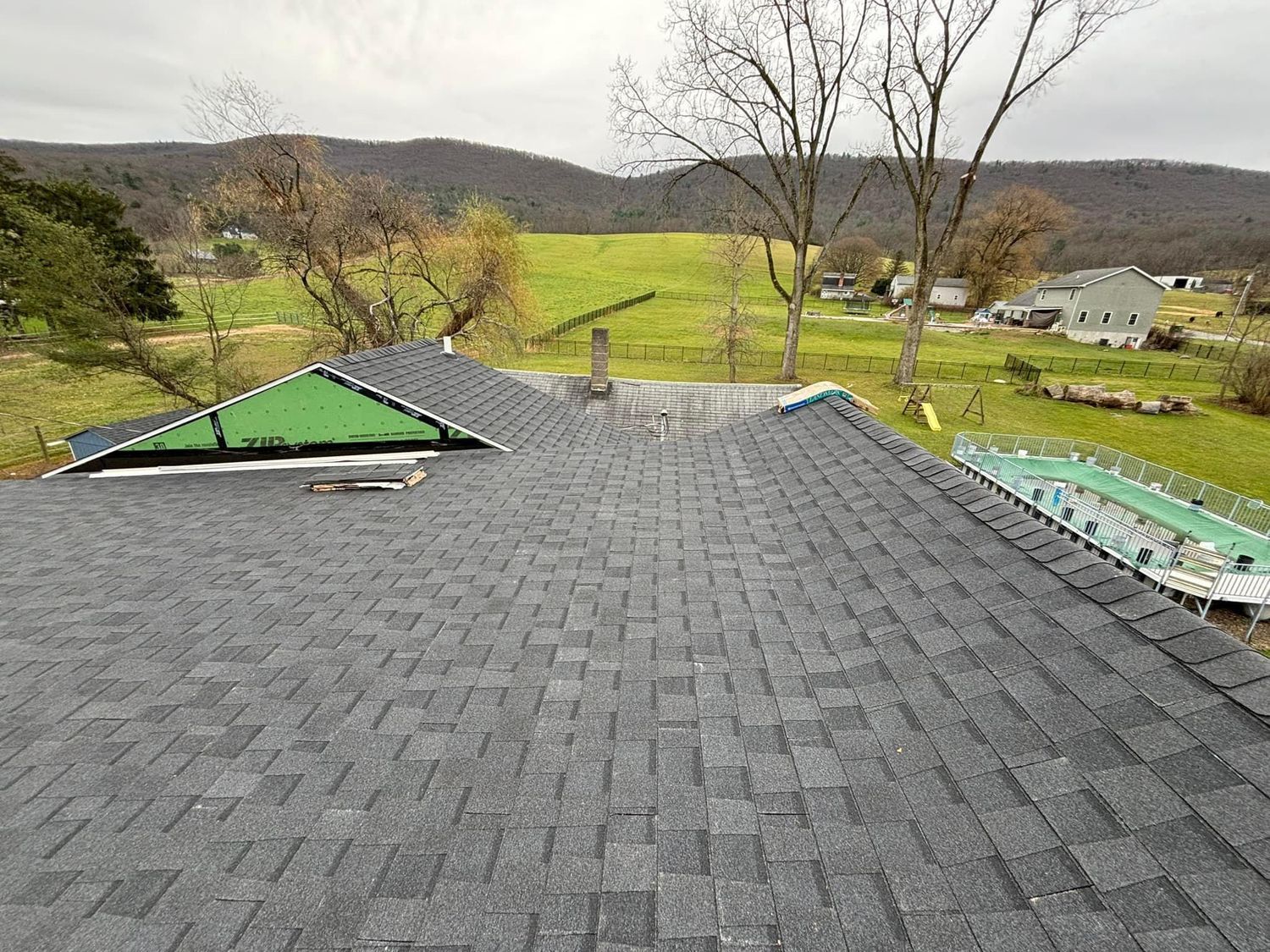 A view from a roof showing new grey shingles, a section with green underlayment, and a yard with a pool in the distance.