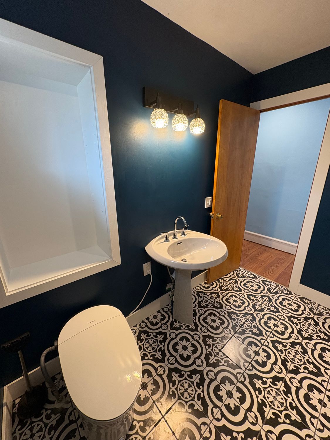 A bathroom with dark blue walls, patterned black and white tile flooring, a pedestal sink, and a wall-mounted light.