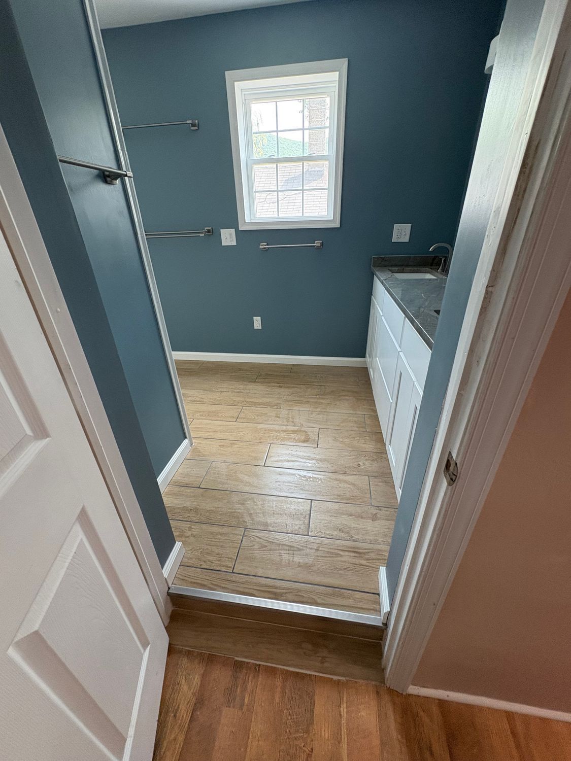 A view through a doorway into a small room with blue walls, a white window, wood-look flooring, and a white vanity.