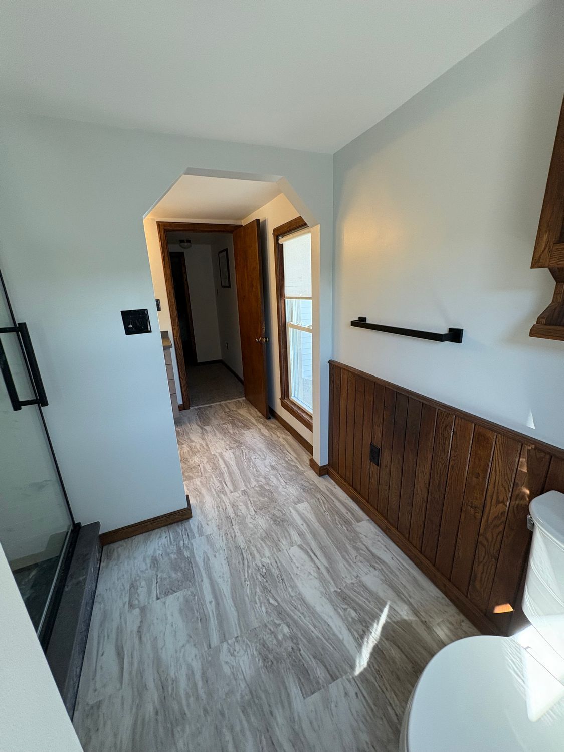 Bathroom with light grey walls, white wood-look flooring, and wood wainscoting leading to a doorway.