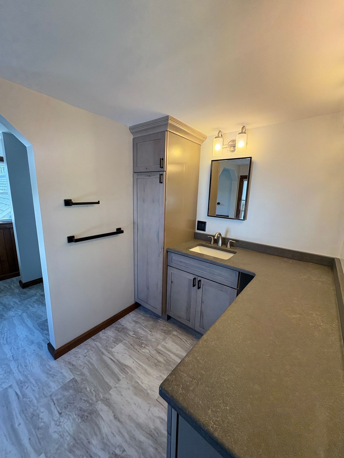 A bathroom with a gray wood-toned vanity, a tall cabinet, stone countertops, a mirror, and two wall-mounted towel bars.