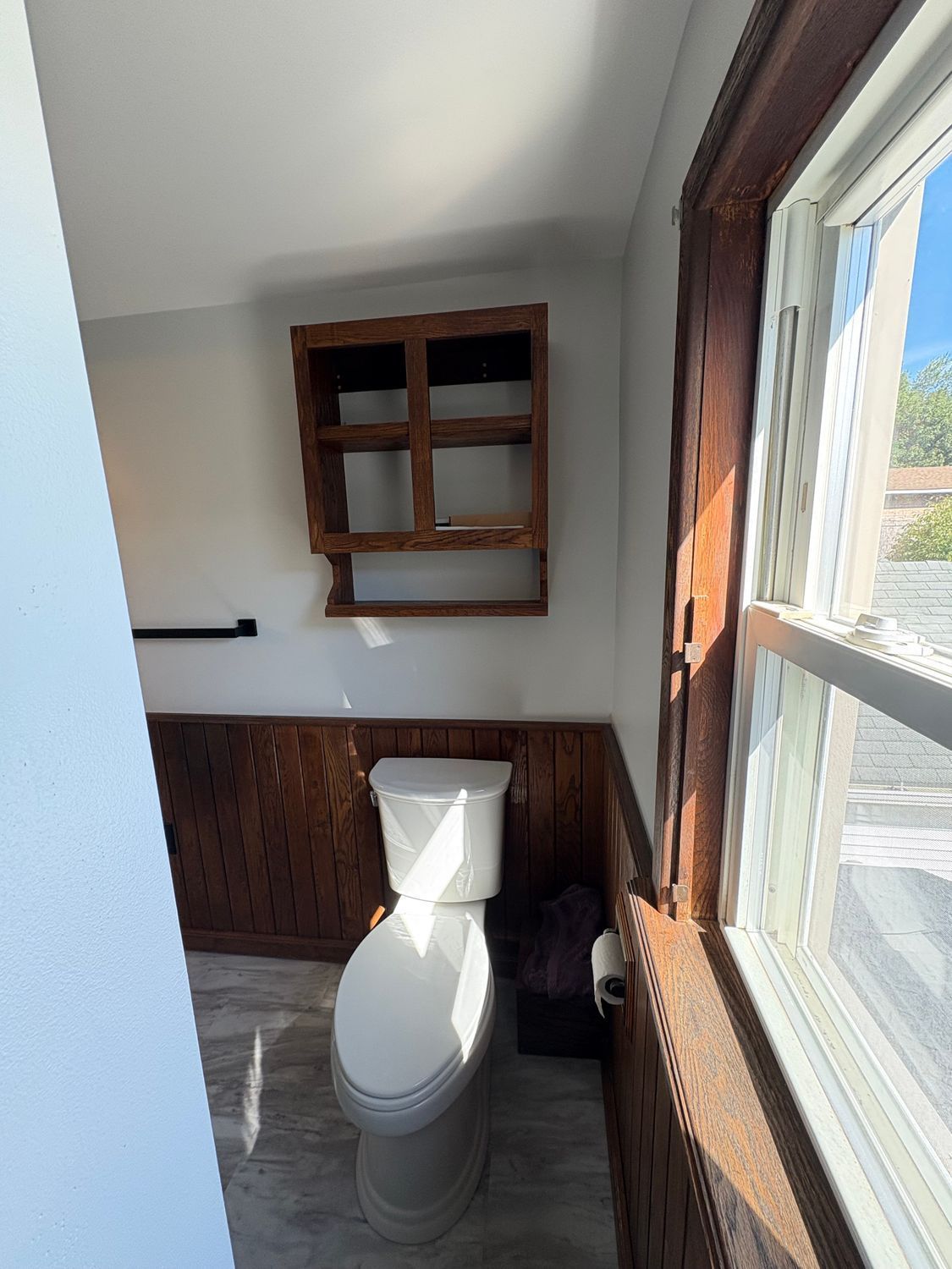 A bathroom corner with a white toilet, wood paneling, wood shelving unit, and a window letting in natural light.