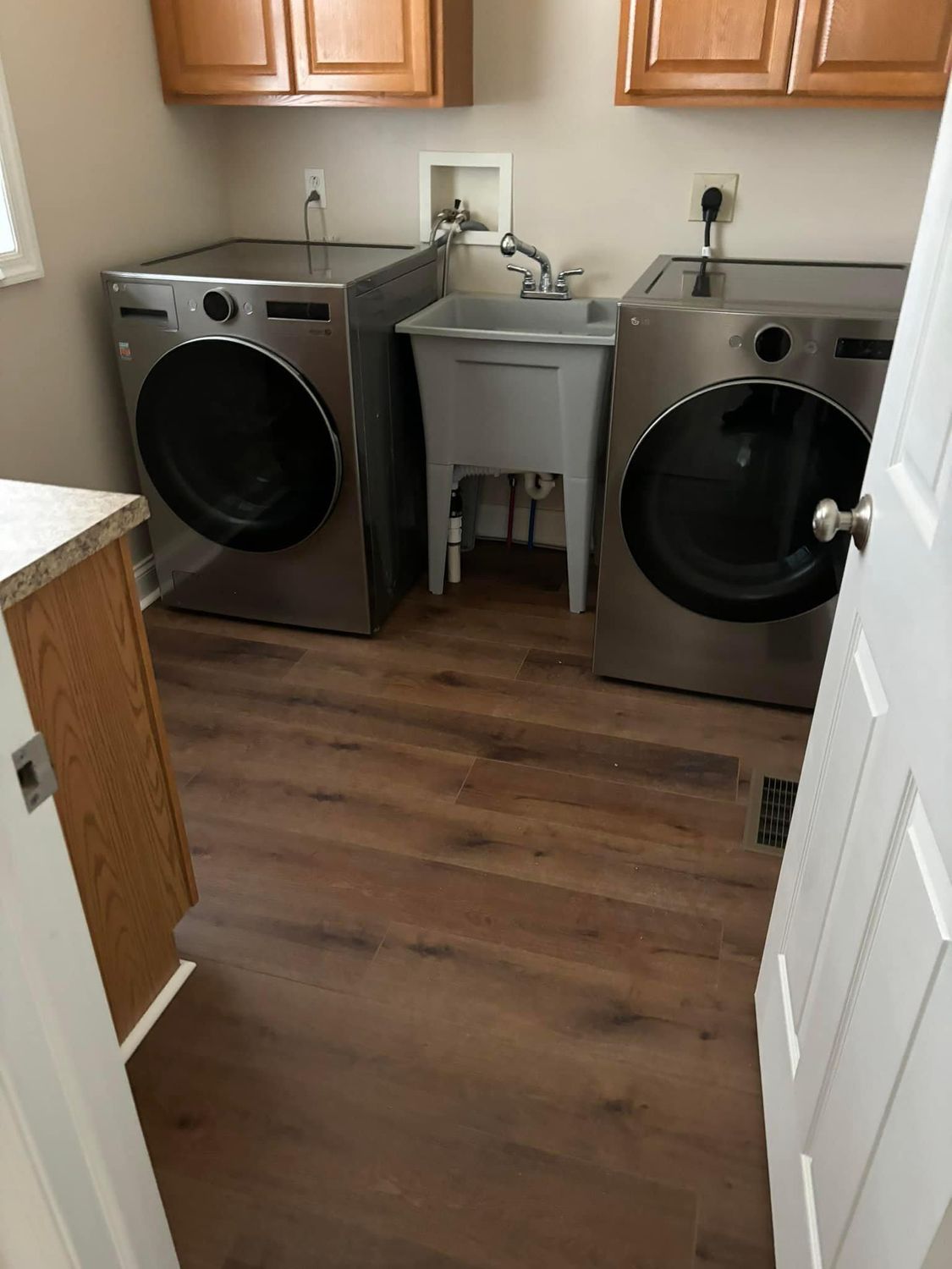 A laundry room with two gray, front-load appliances and a utility sink between them, set against wood cabinets and floor.