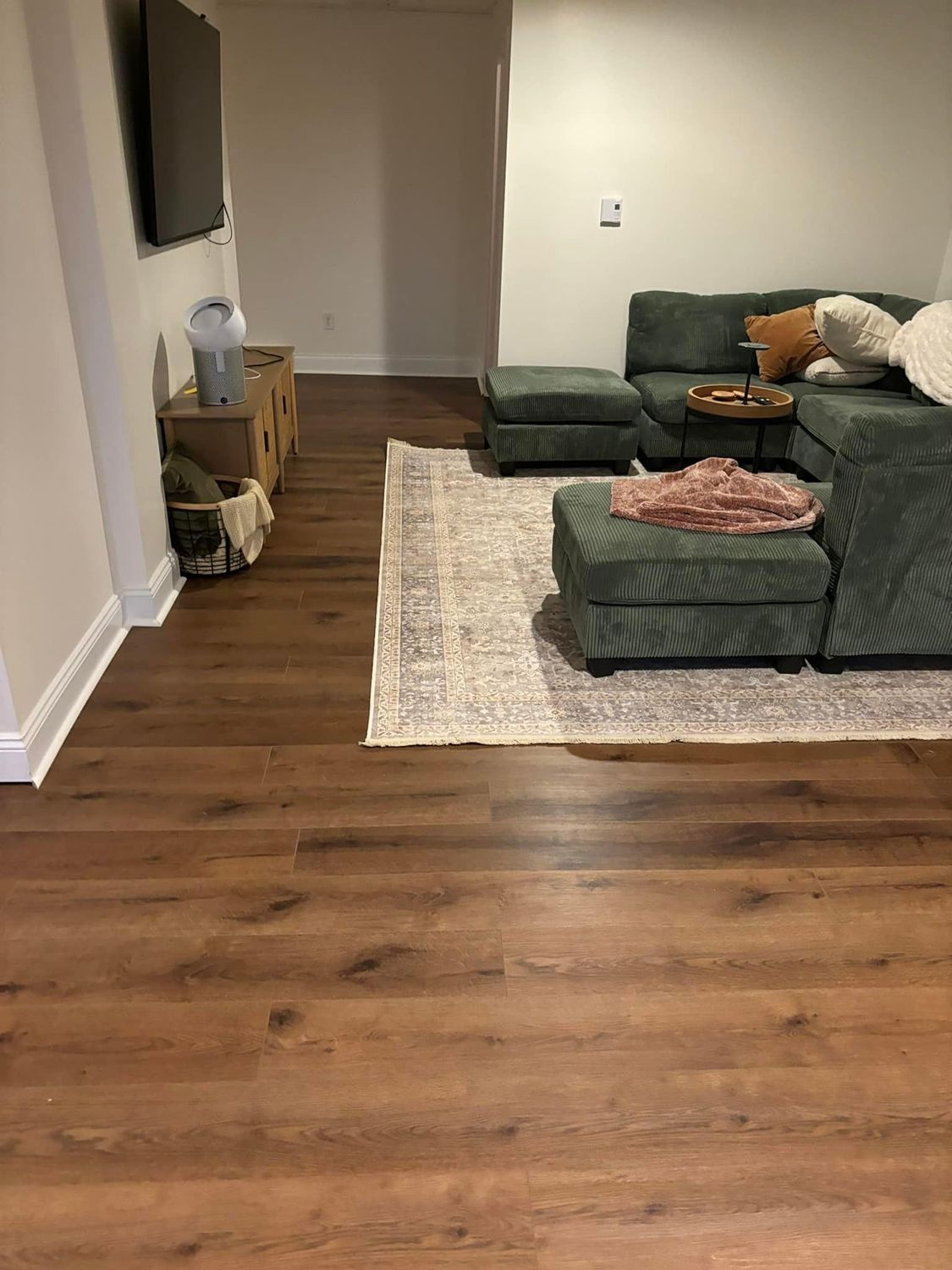 A living room with wood-look flooring, a green sectional sofa, a rug, and a small side table holding a lamp.