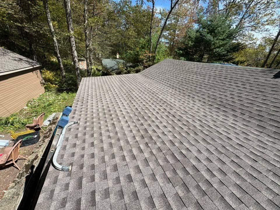 A high-angle view of a gray shingled roof under a clear blue sky, next to a brick house and green trees.