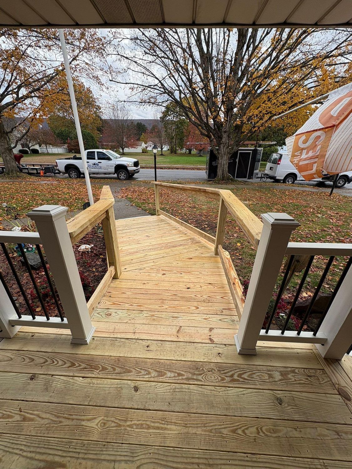 A wooden ramp with railings extends from a porch down toward a residential street with parked vehicles and fall trees.