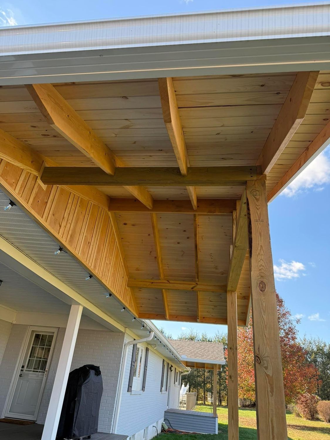 A wooden porch roof addition under construction, attached to a house with white siding under a clear blue sky.