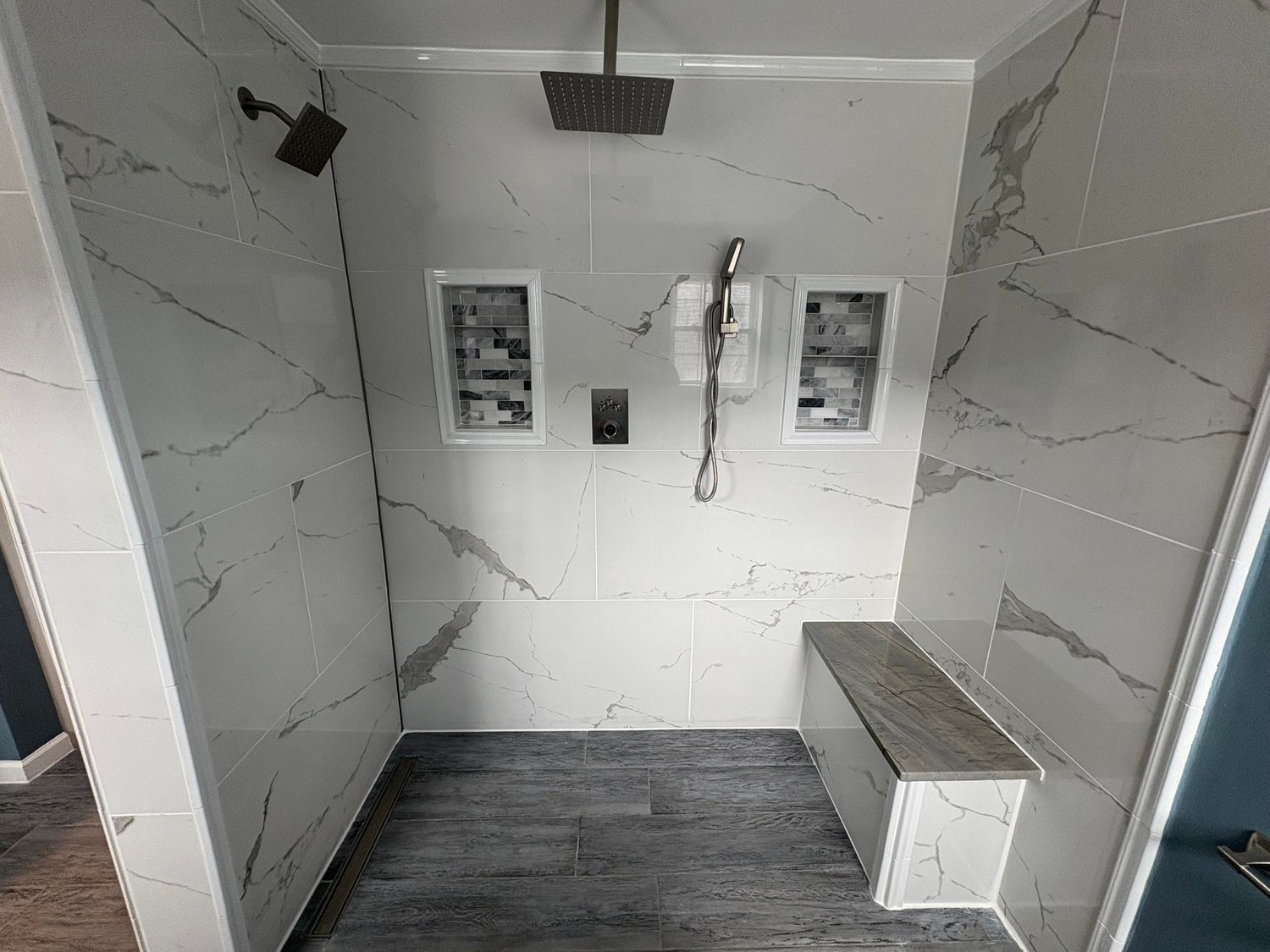A modern shower with white marble-veined tile walls, a ceiling rain showerhead, wall fixtures, two niches, and a bench.
