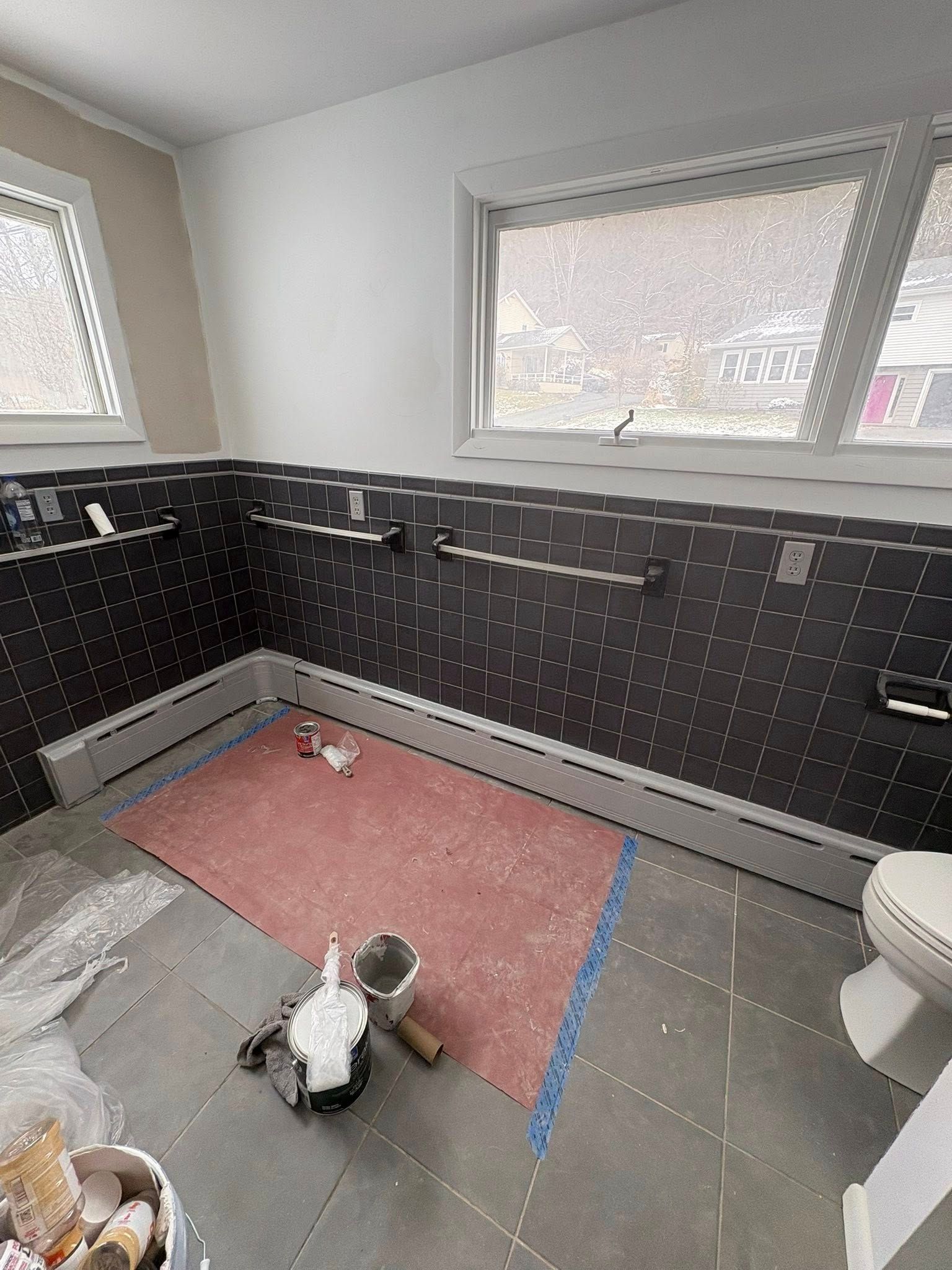 A bathroom under renovation with black-tiled walls, gray floor tiles, a pink protective mat, and painting supplies.