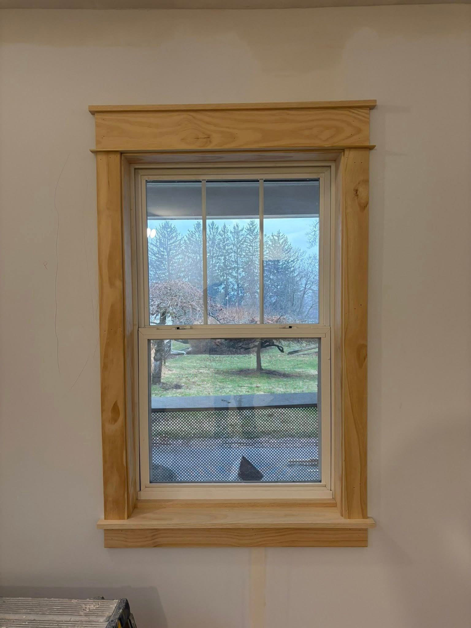 A window with unfinished light-colored wood trim against a plain white wall, overlooking a yard with a lattice fence.