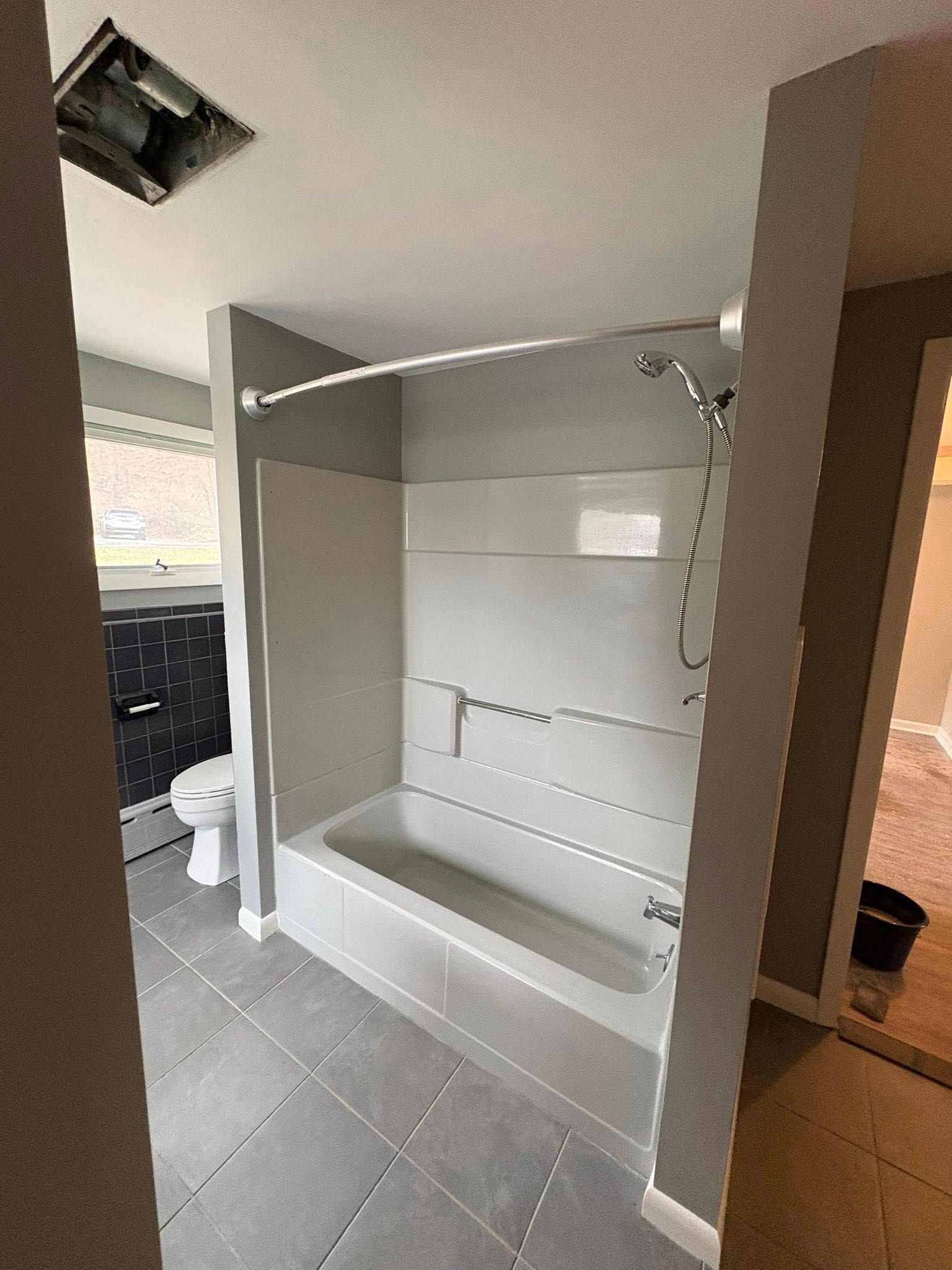 A bathroom featuring a white bathtub with a showerhead, gray tiled floors, and a wall-mounted toilet in the background.
