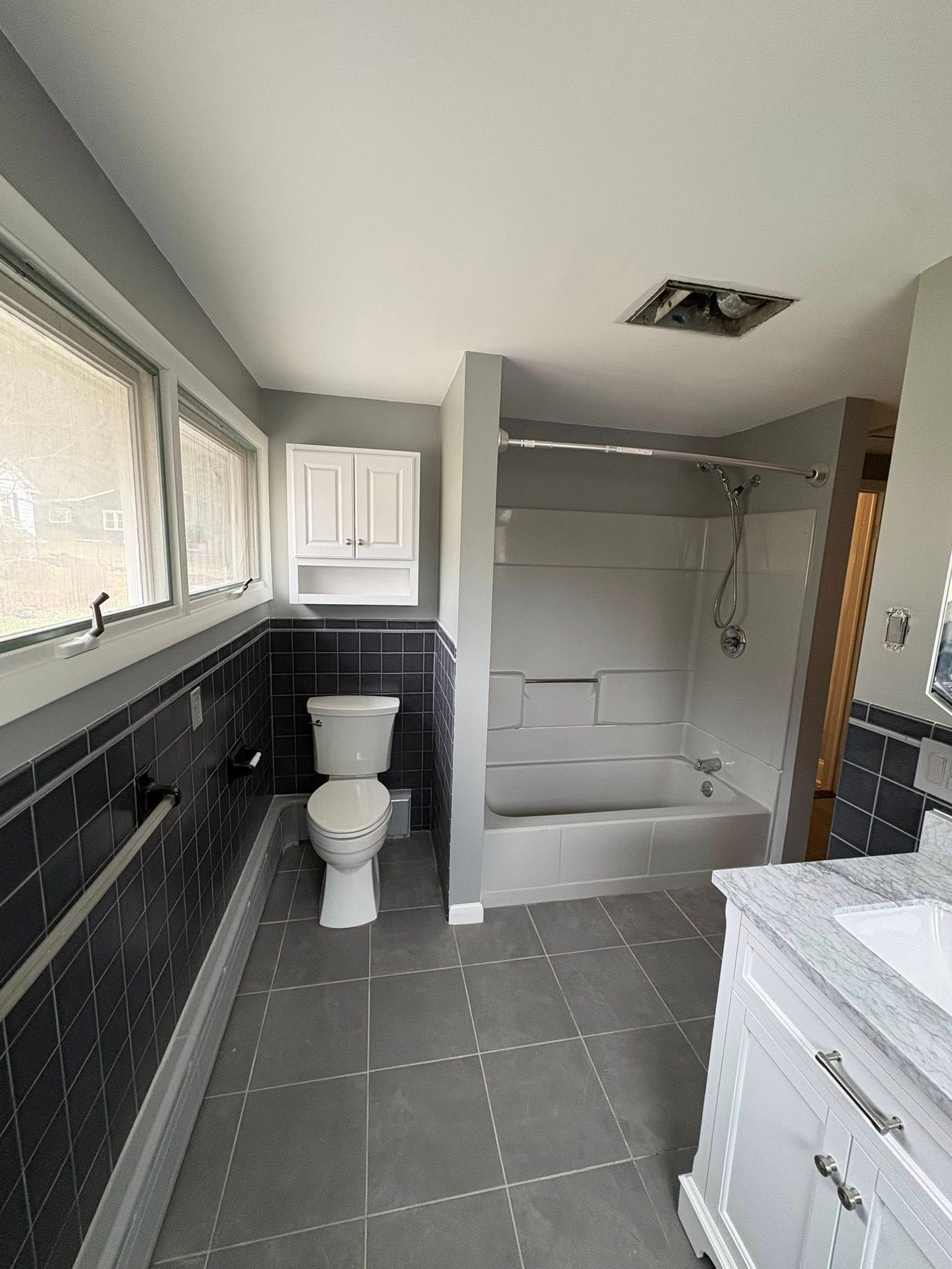 A bright bathroom with dark grey tiled walls, a white toilet, and a white shower-tub combination.