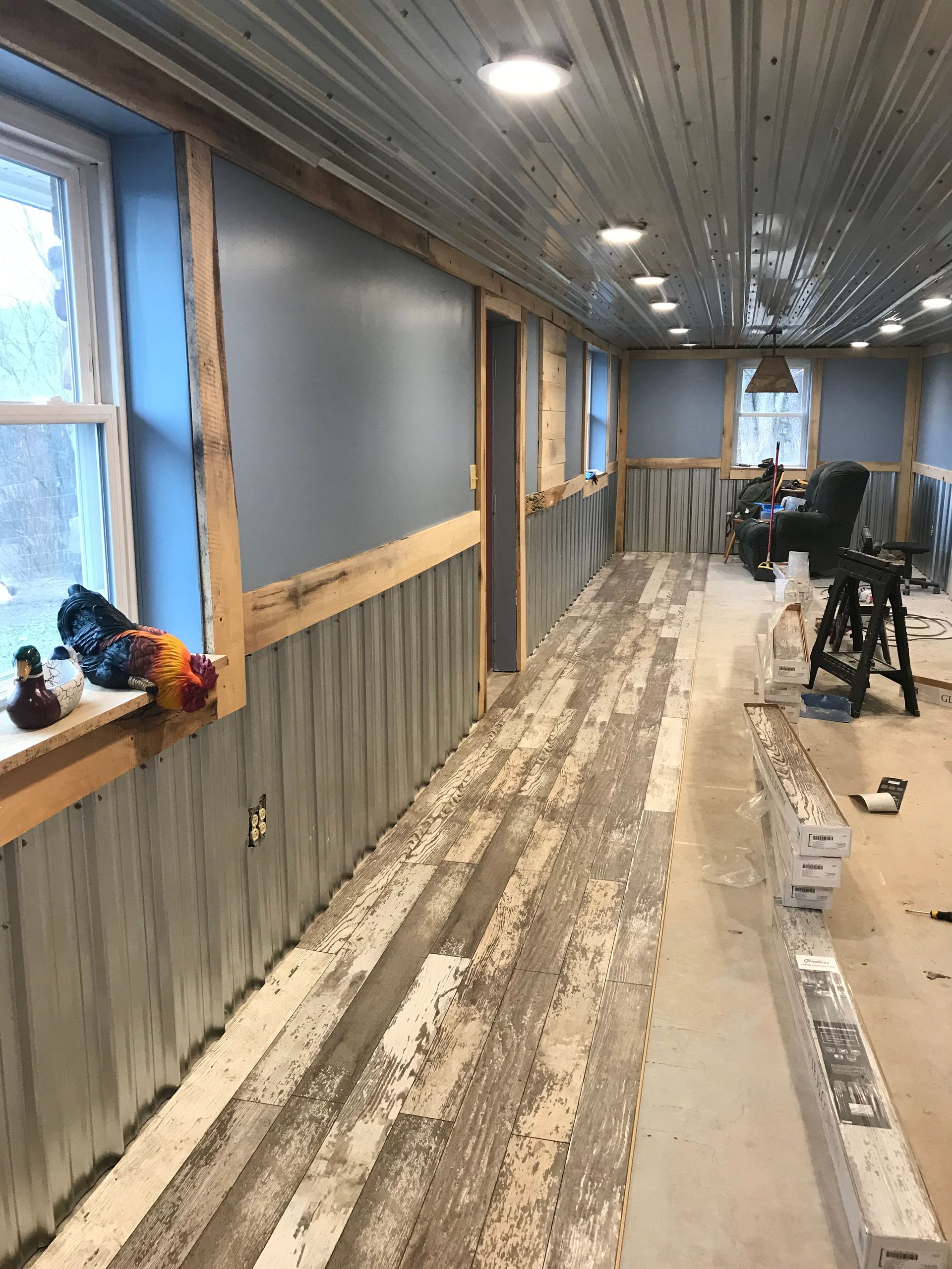 A hallway under renovation with corrugated metal ceilings, wainscoting, grey walls, and exposed subflooring.