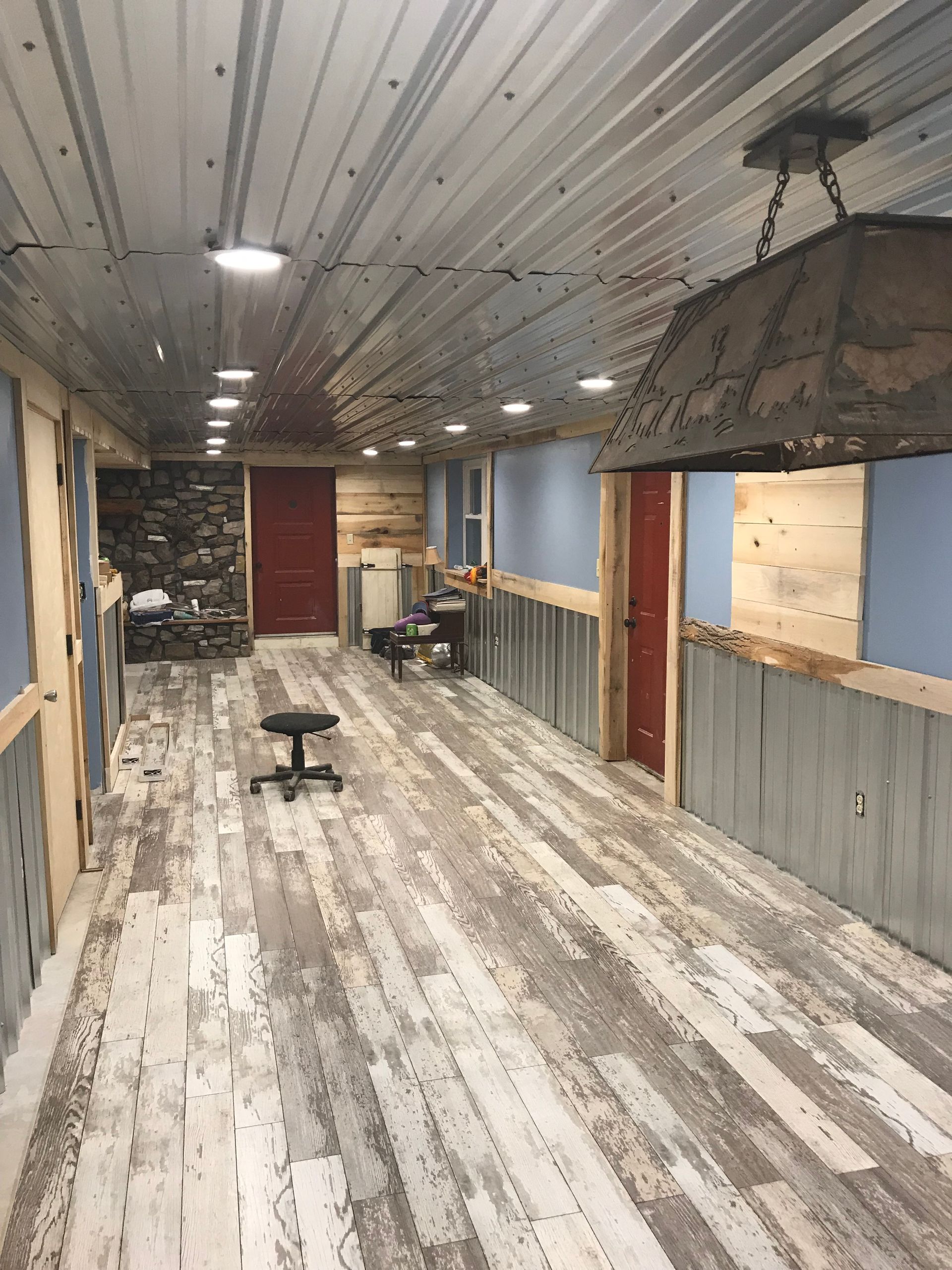 A long, rustic room under renovation with a corrugated metal ceiling, wood plank walls, and weathered wood-look flooring.