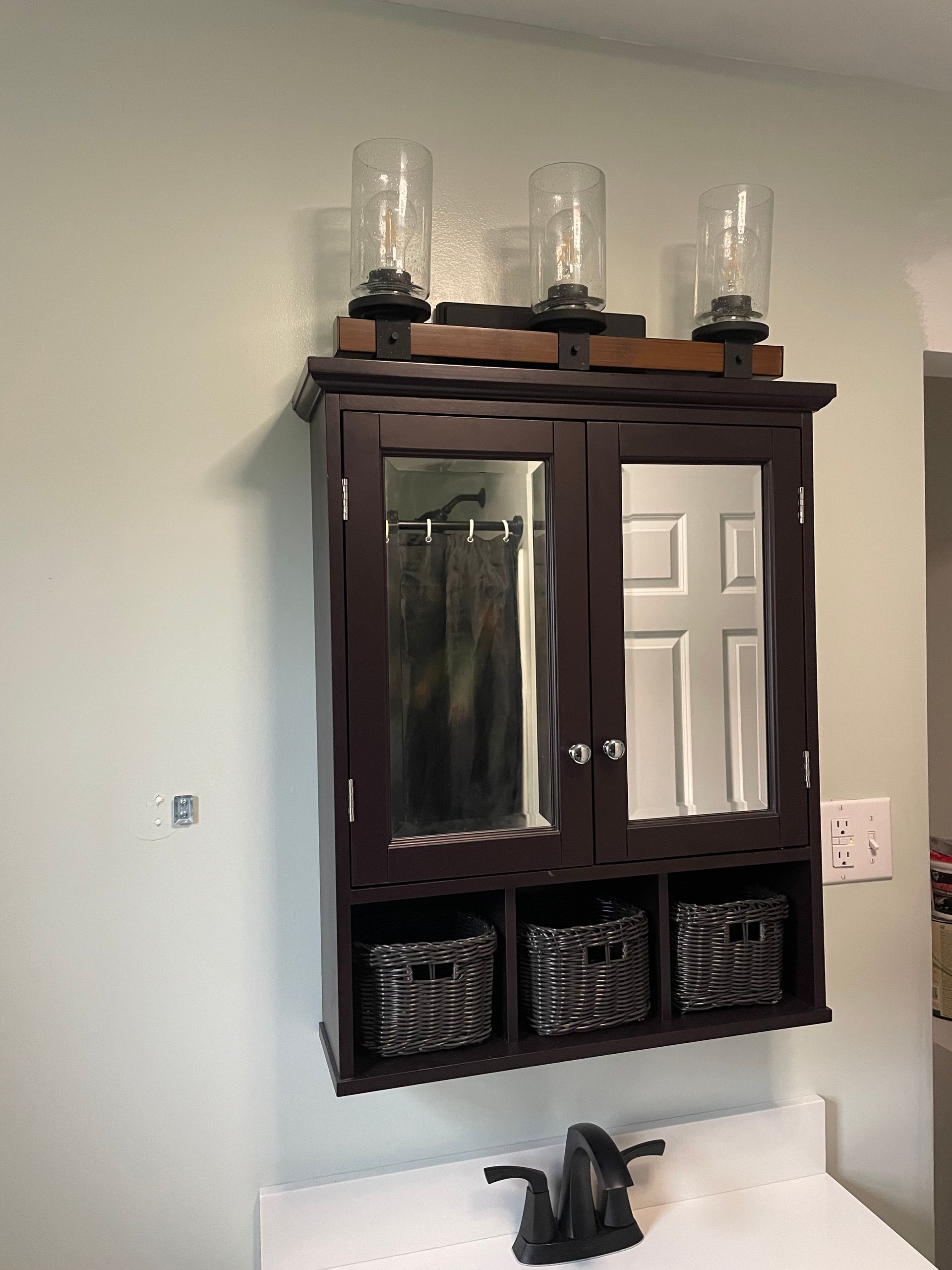 A dark wood bathroom medicine cabinet with mirrored doors, open storage cubbies with baskets, and three glass light fixtures.