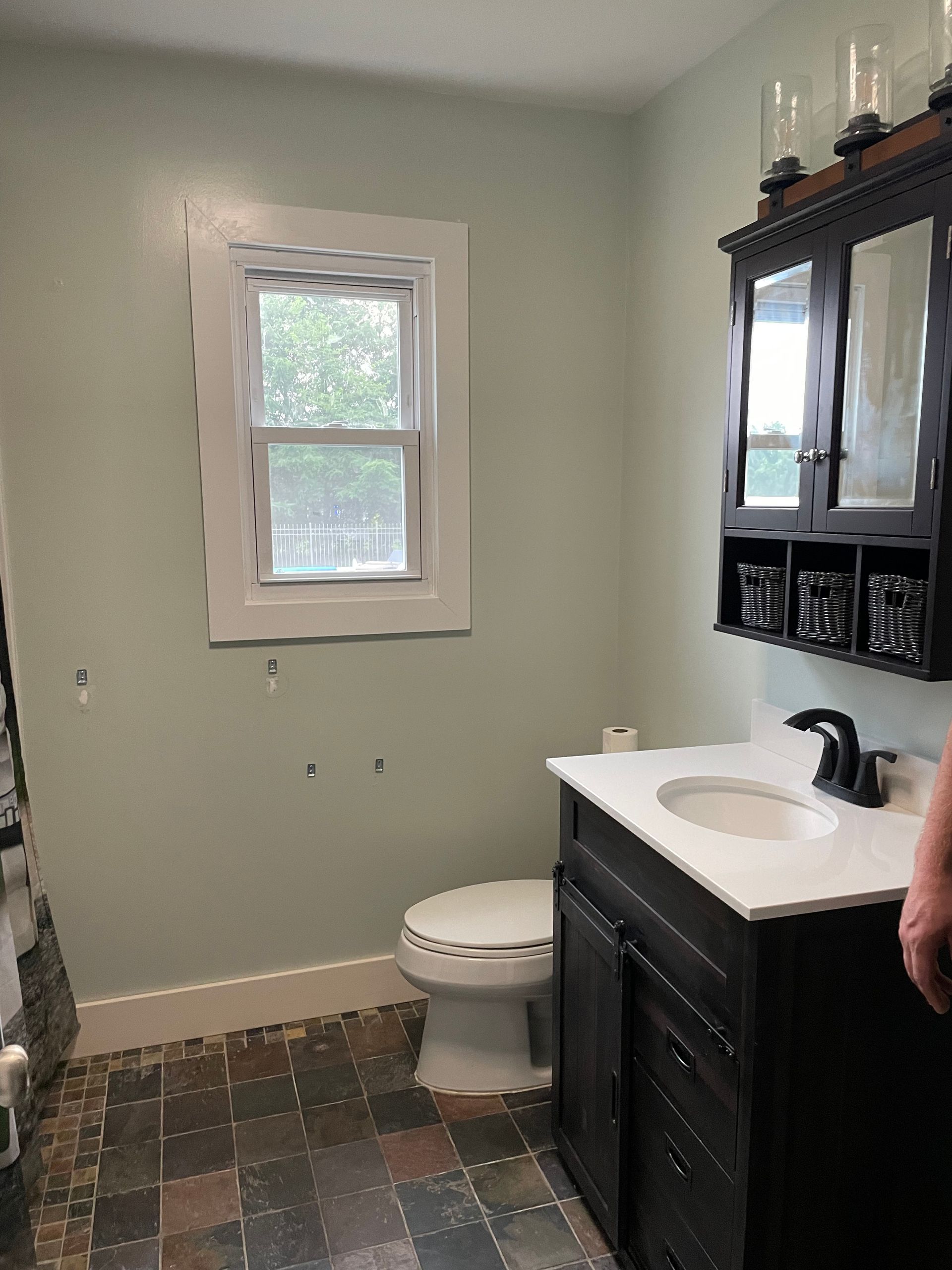 A bathroom with light green walls, a black vanity, a matching wall cabinet, a window, and a white toilet.