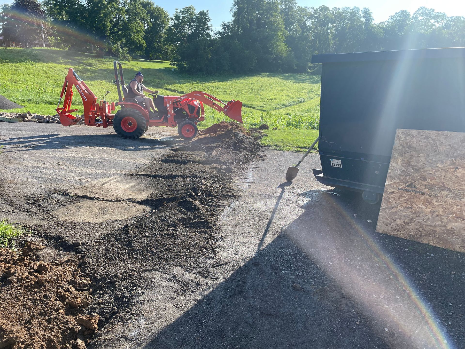 An orange tractor with a backhoe attachment works on a gravel driveway near a black dumpster on a sunny day.