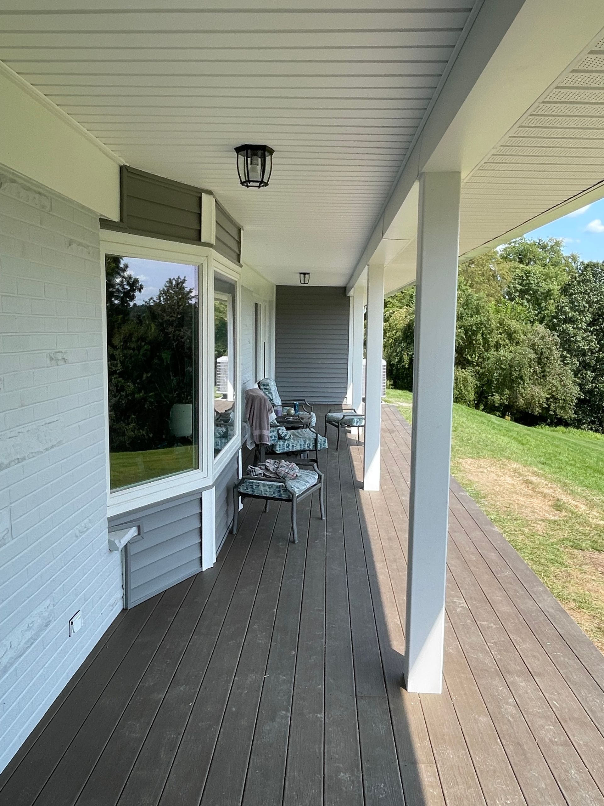 A long, covered outdoor deck with gray wooden flooring, white support pillars, and a light gray brick wall with windows.