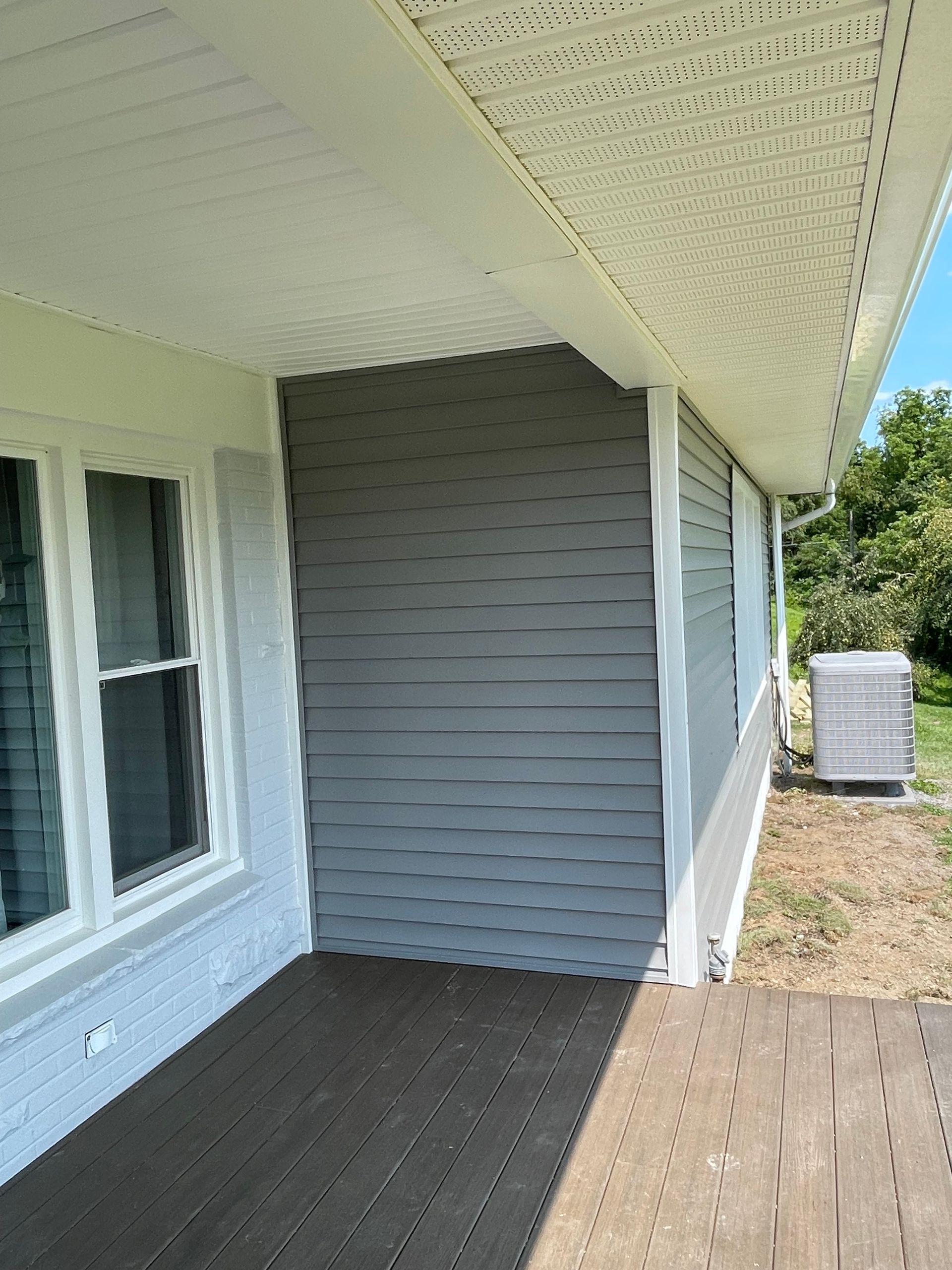 A grey-sided corner of a house exterior with a white window, a white soffit overhang, and a brown wooden deck.