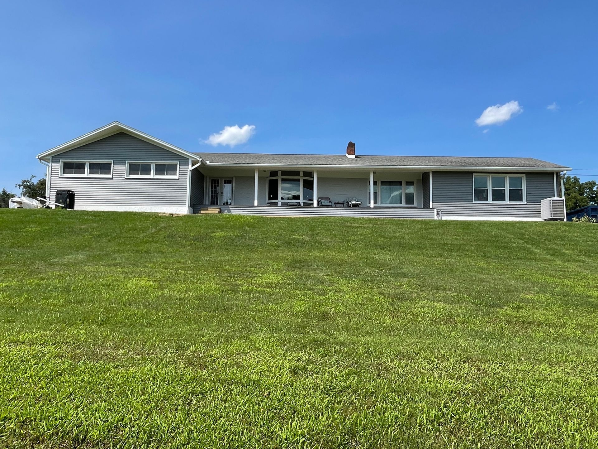 A one-story gray house with a gable roof sits atop a large, green grassy hill under a bright blue sky.