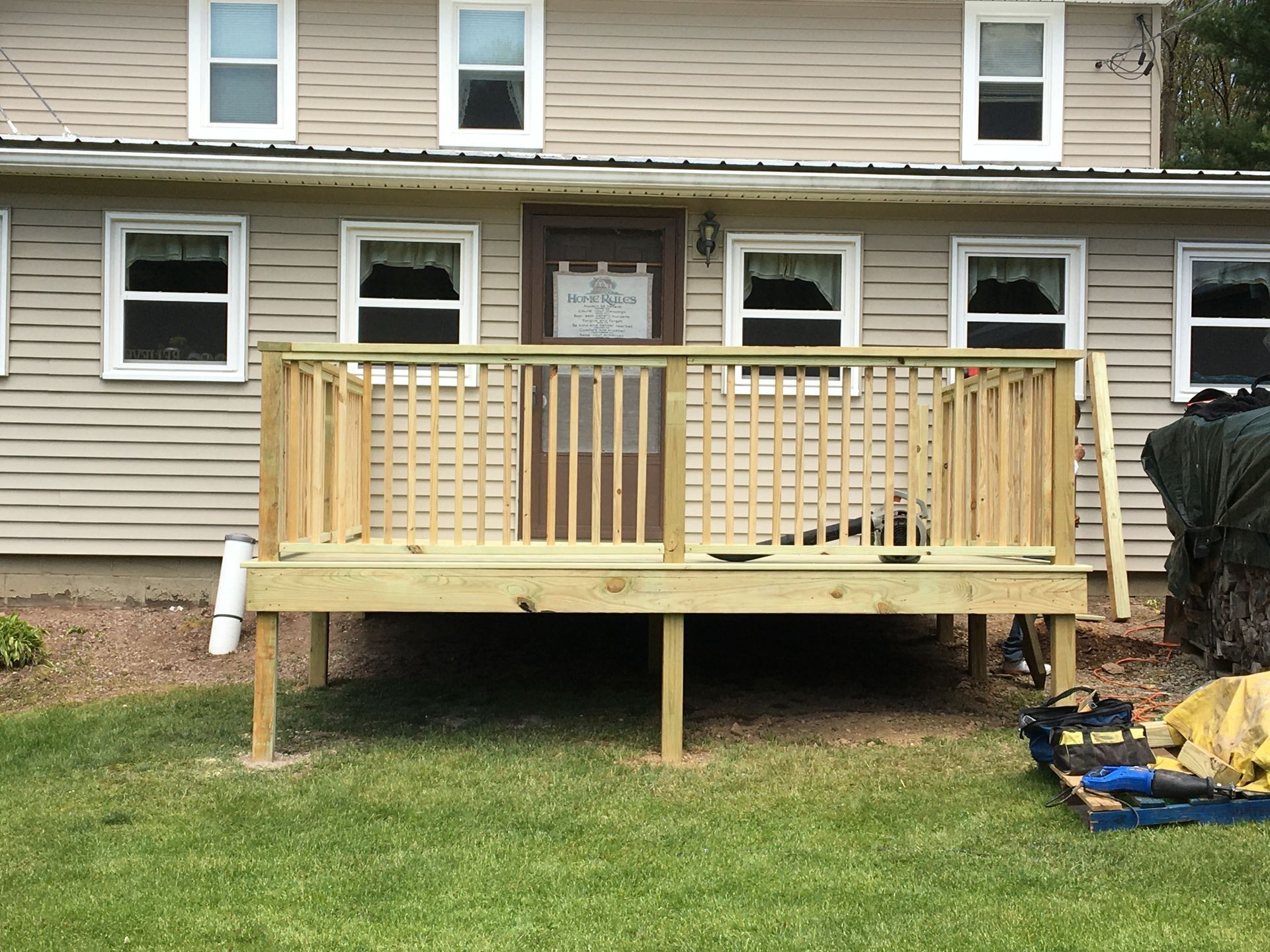 A newly constructed wooden deck with railings attached to the back of a light brown siding house on a grassy lawn.
