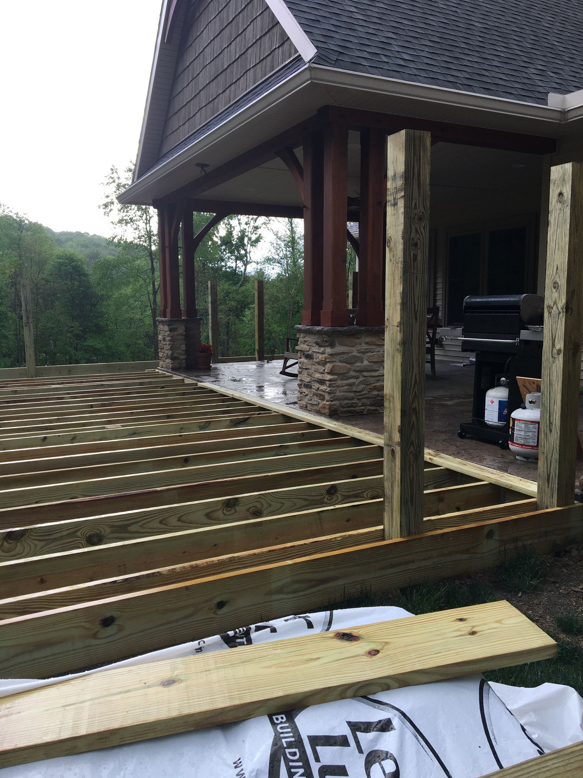 Wooden framing for a new deck under construction, positioned beside a covered stone and timber porch.