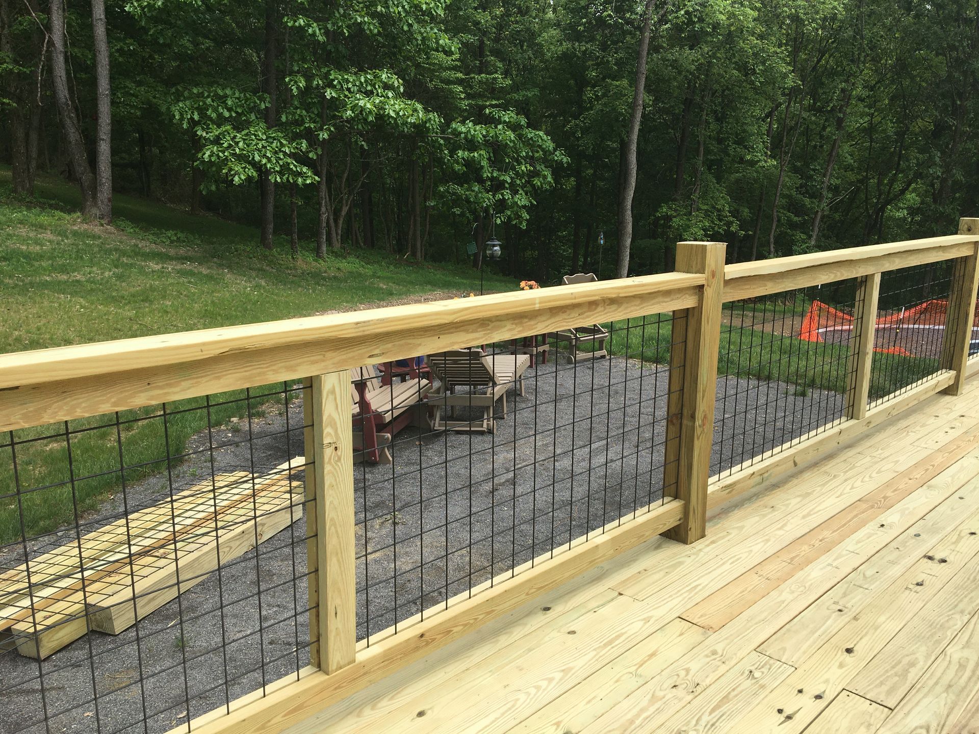 A wooden deck railing with a wire mesh panel insert, overlooking a gravel area and a wooded backyard.
