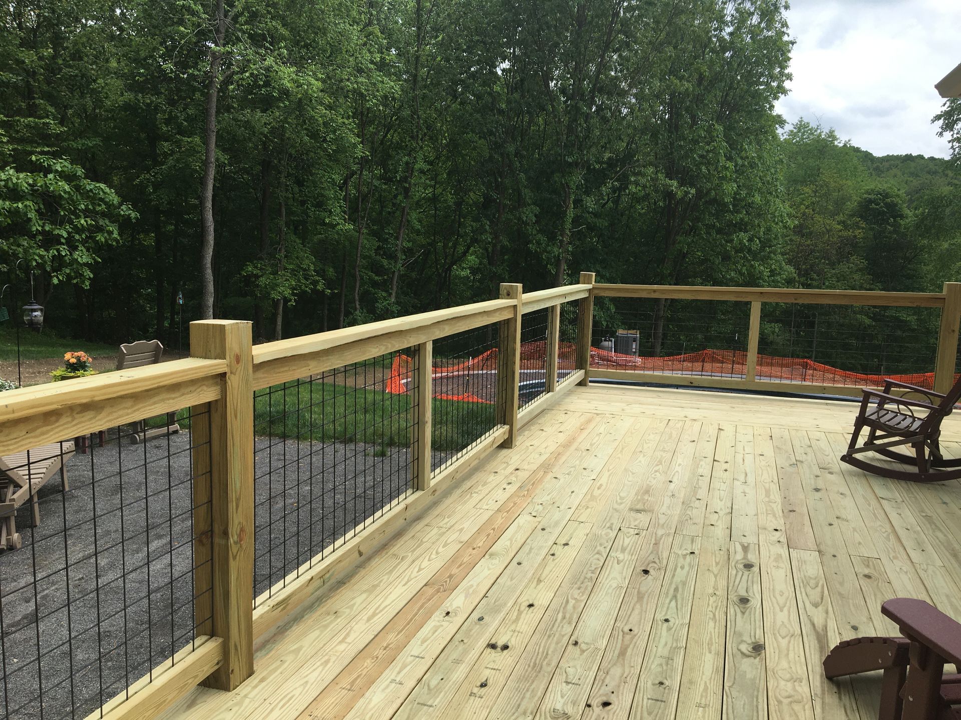 A wooden deck with a railing featuring metal cable infill, overlooking a grassy area, trees, and construction fencing.