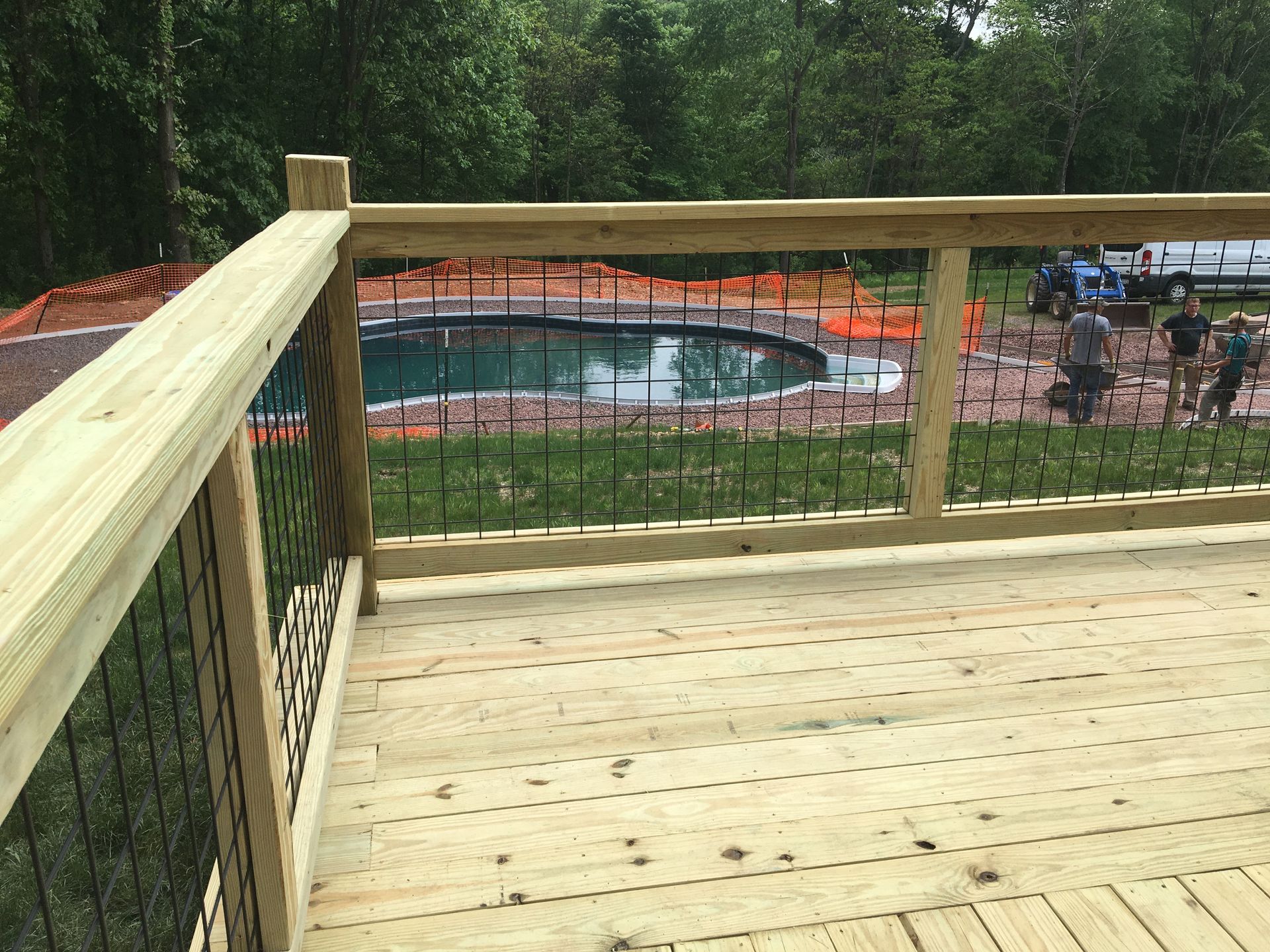 A view from a wooden deck looking out toward an outdoor pool under construction, surrounded by orange safety fencing.