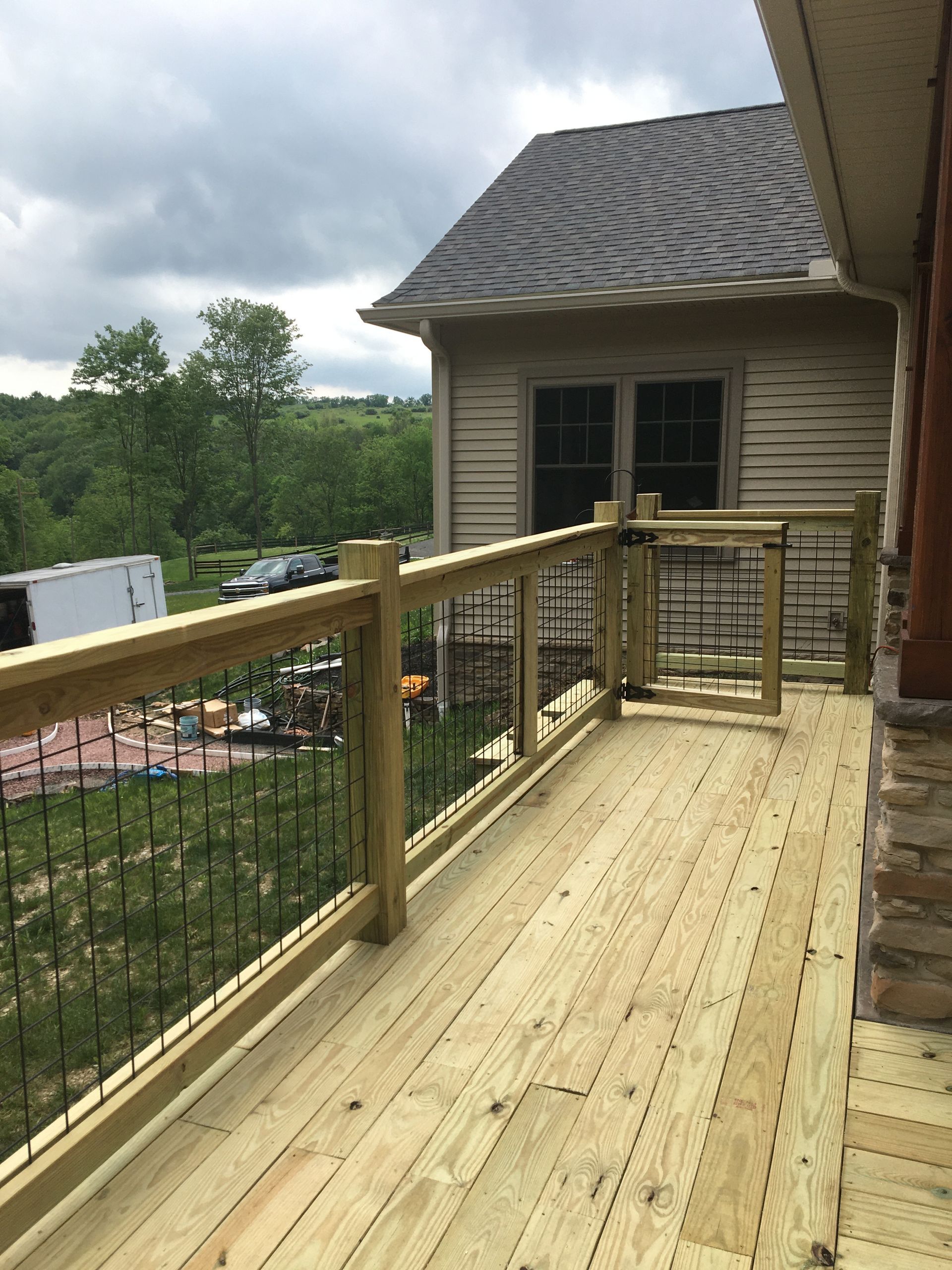 A wooden deck with a railing and metal balusters overlooking a grassy yard, a tree line, and a neighboring house.