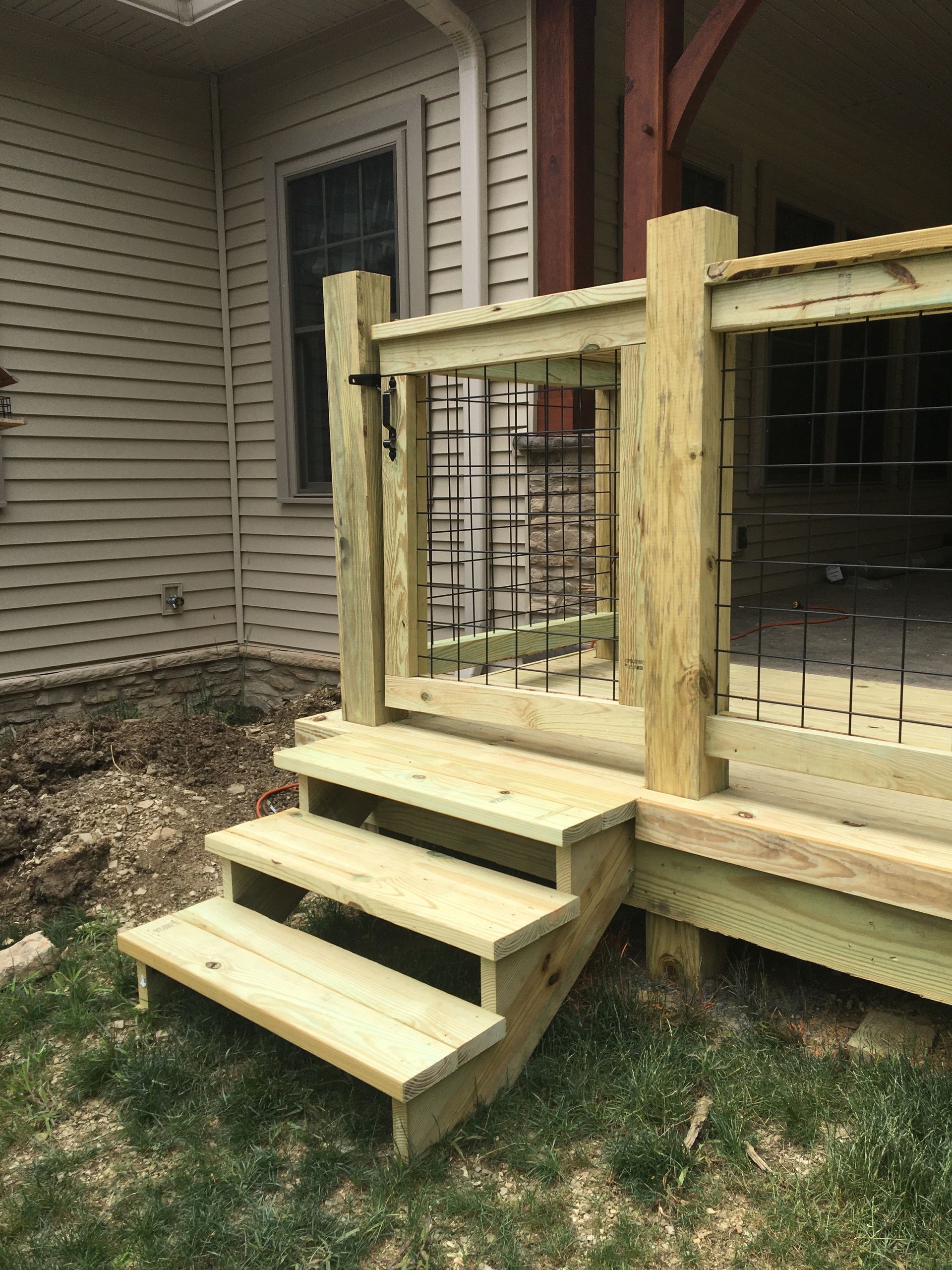 Three wooden steps lead up to a deck with a cable railing and a gate next to a house with beige siding.