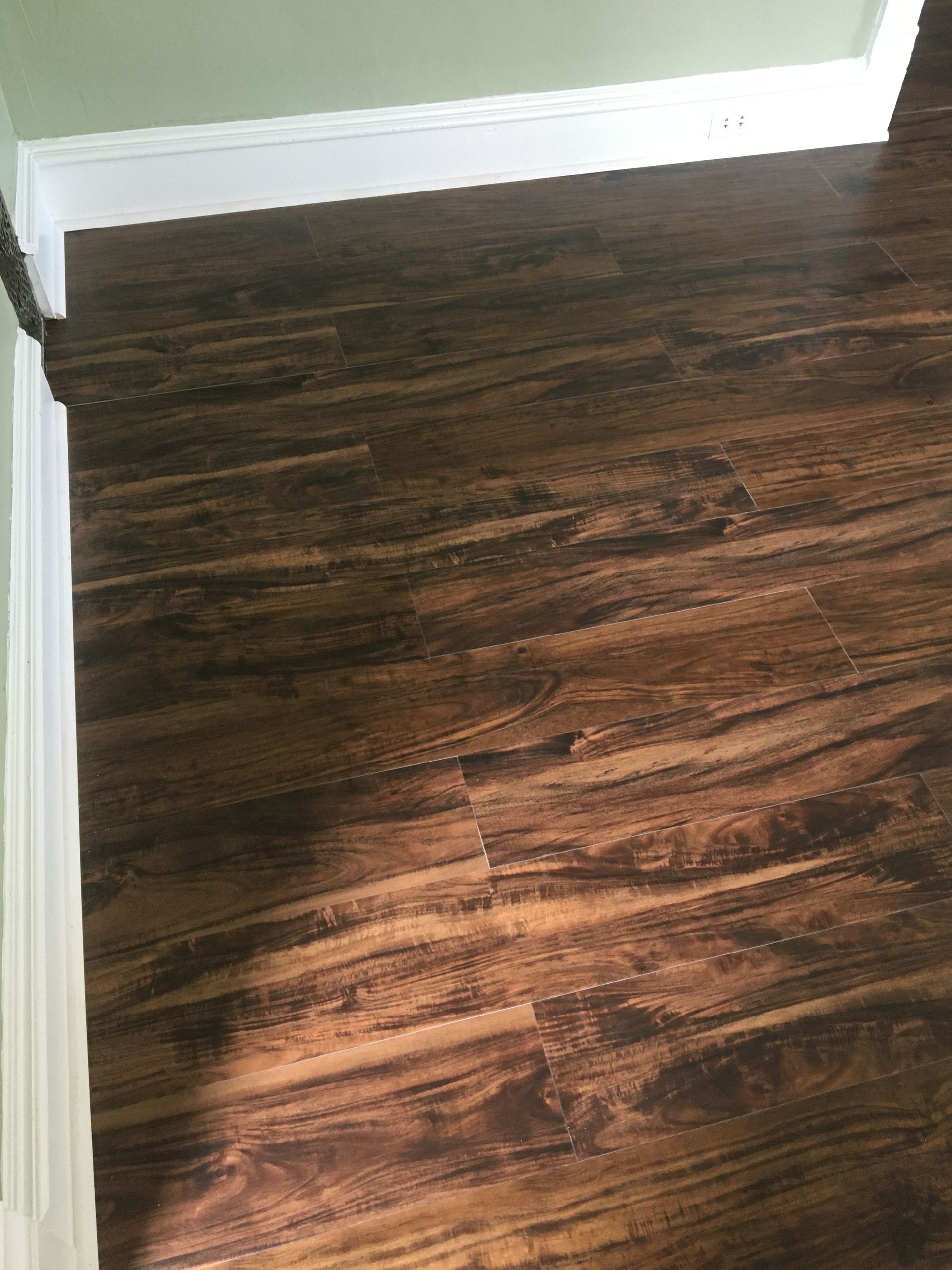 Dark brown wood-grain laminate flooring meeting white baseboards against a sage green wall.
