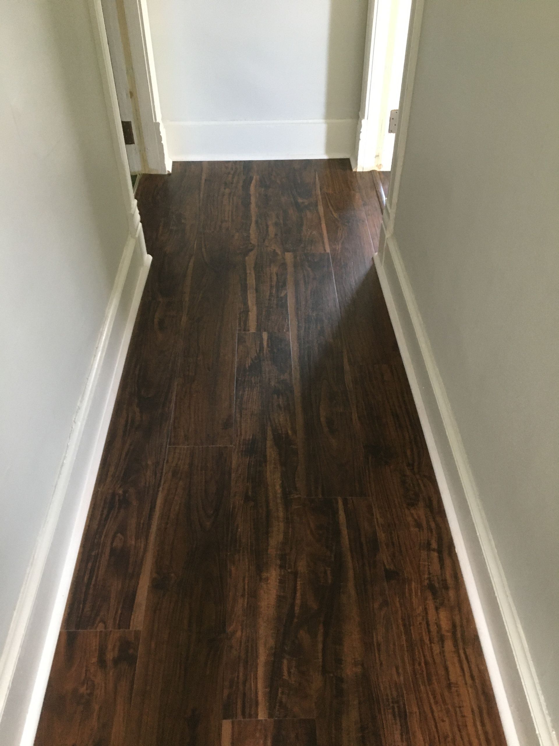 A narrow hallway with dark wood-look plank flooring and white baseboards, leading to a doorway.