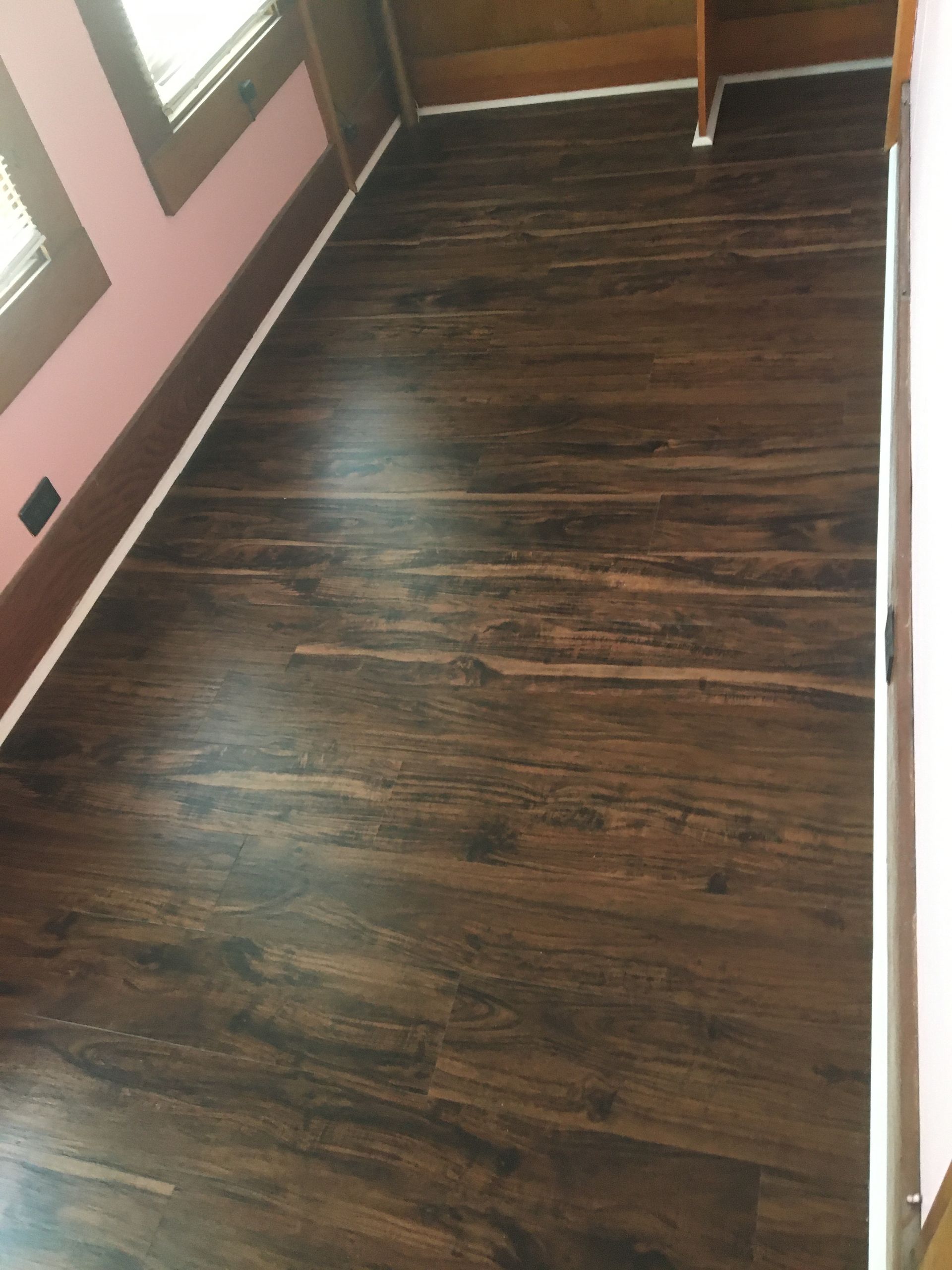 Dark wood-look laminate flooring installed in a room with light pink walls and white baseboards.