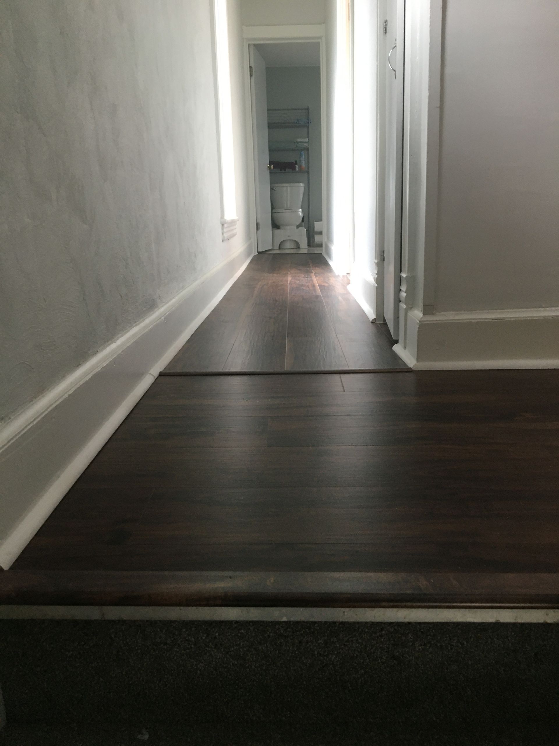 A narrow hallway with dark wood-look flooring leading toward a bathroom with a white toilet at the far end.