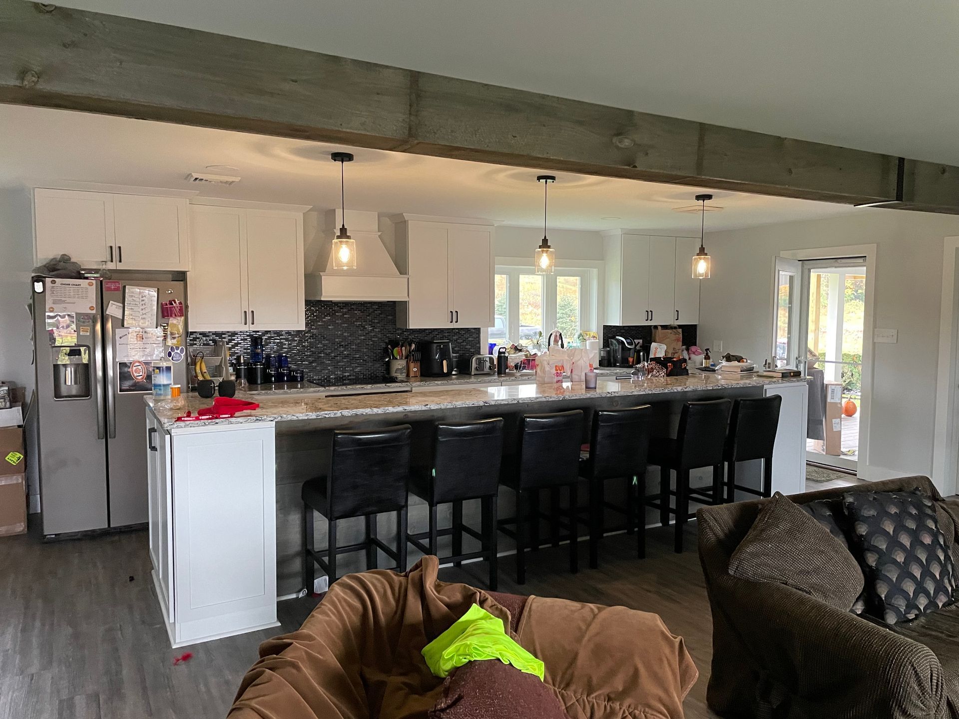 A wide shot of a bright, modern kitchen with white cabinets, a large island with dark stools, and a wooden ceiling beam.