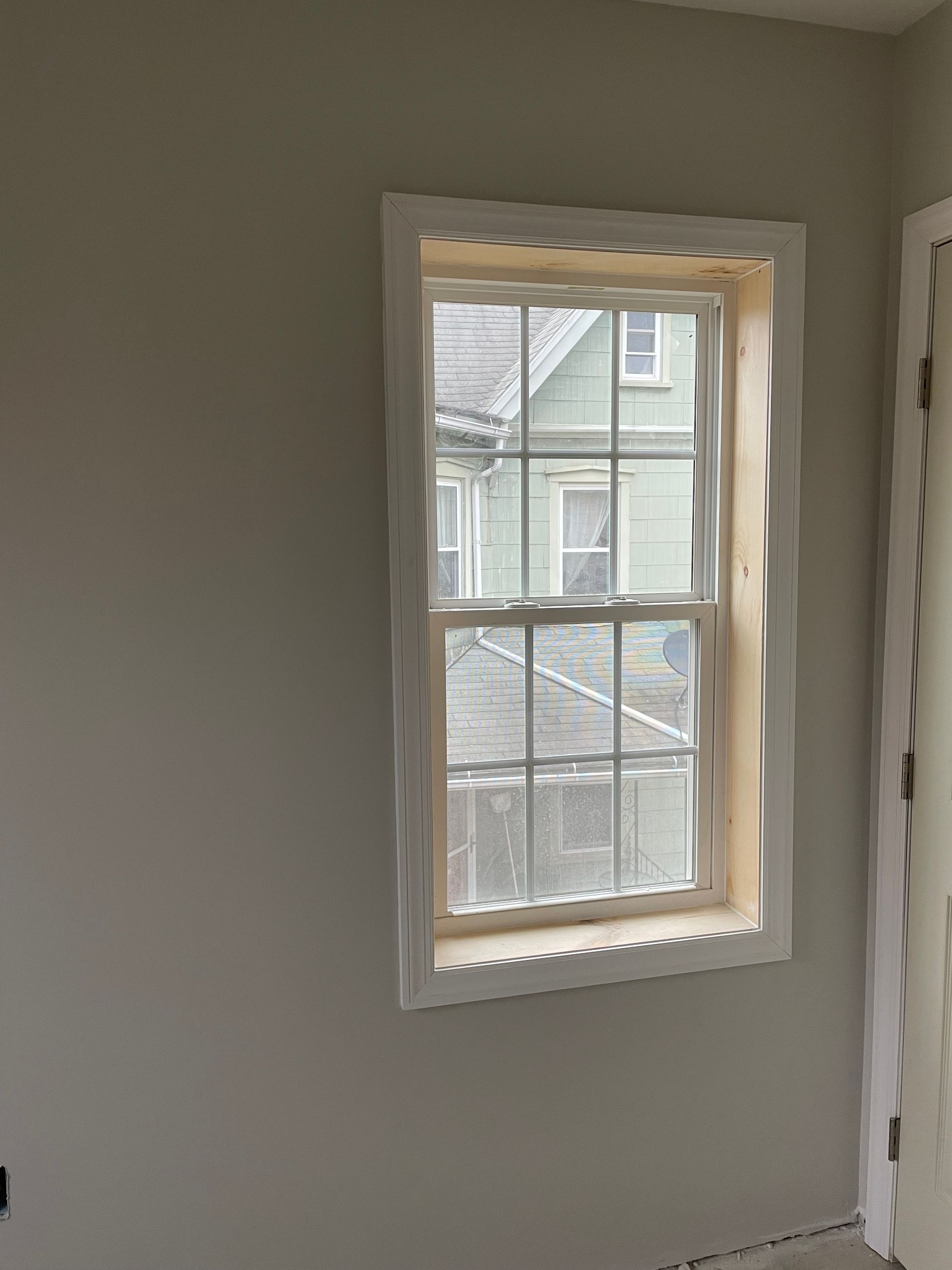 A window with white casing installed on a gray interior wall, showing a view of a neighboring house.