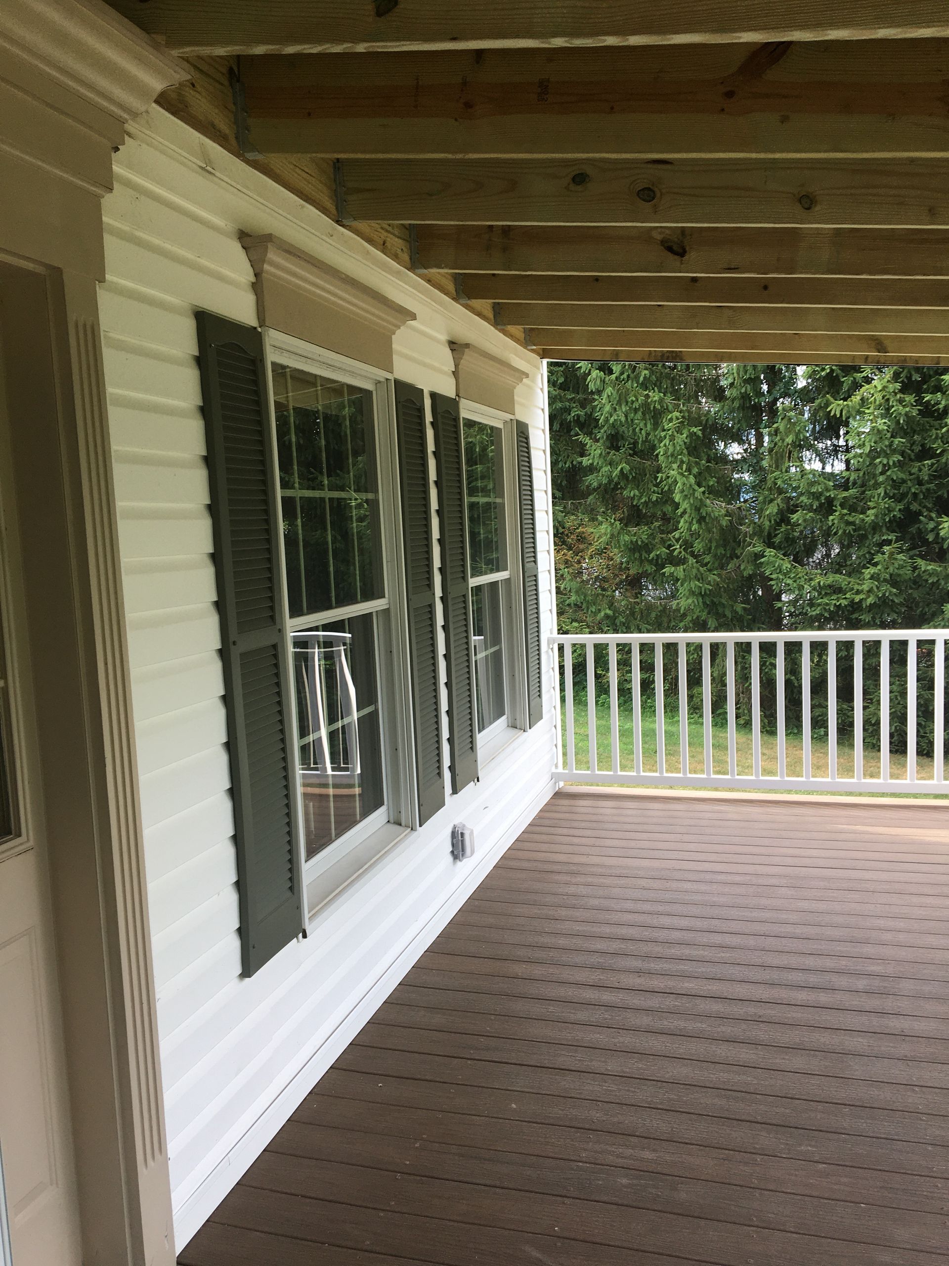 A side view of a porch with white siding, dark window shutters, and a wooden railing looking out at green trees.
