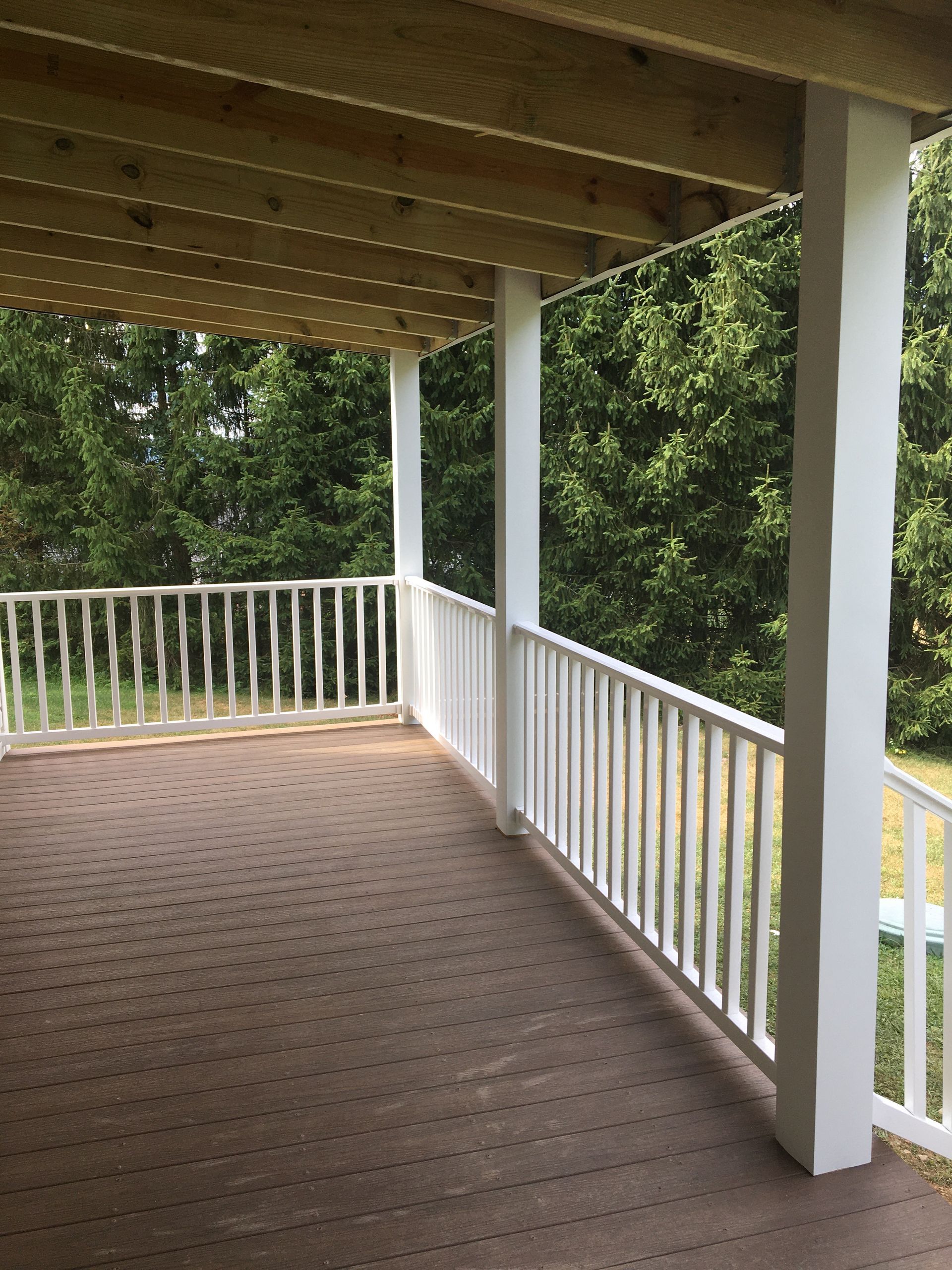 A wooden deck with white railings and support posts, overlooking a line of green trees.