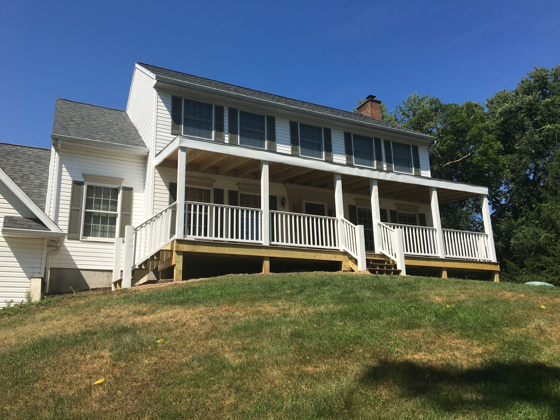 A white two-story house with a new wooden wraparound porch and railing, set on a grassy hill under a clear blue sky.