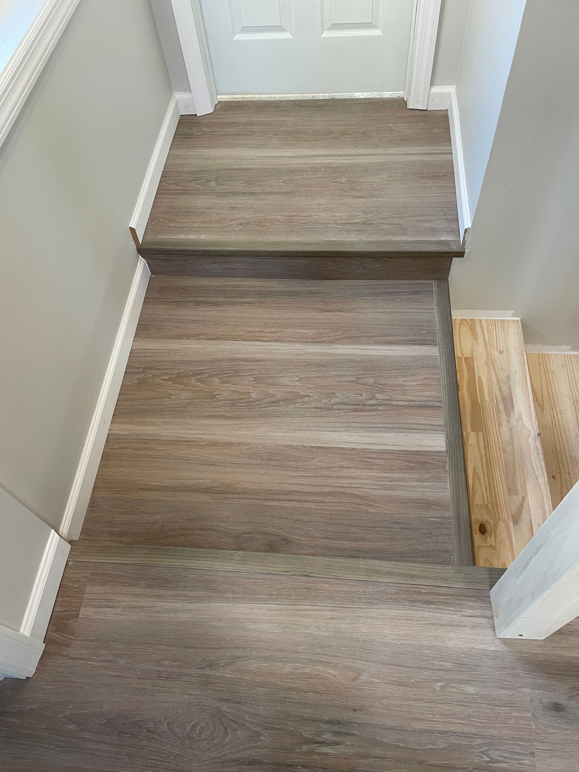 Top-down view of stairs with brown wood-look vinyl flooring leading to a landing, with unfinished wood stairs to the right.